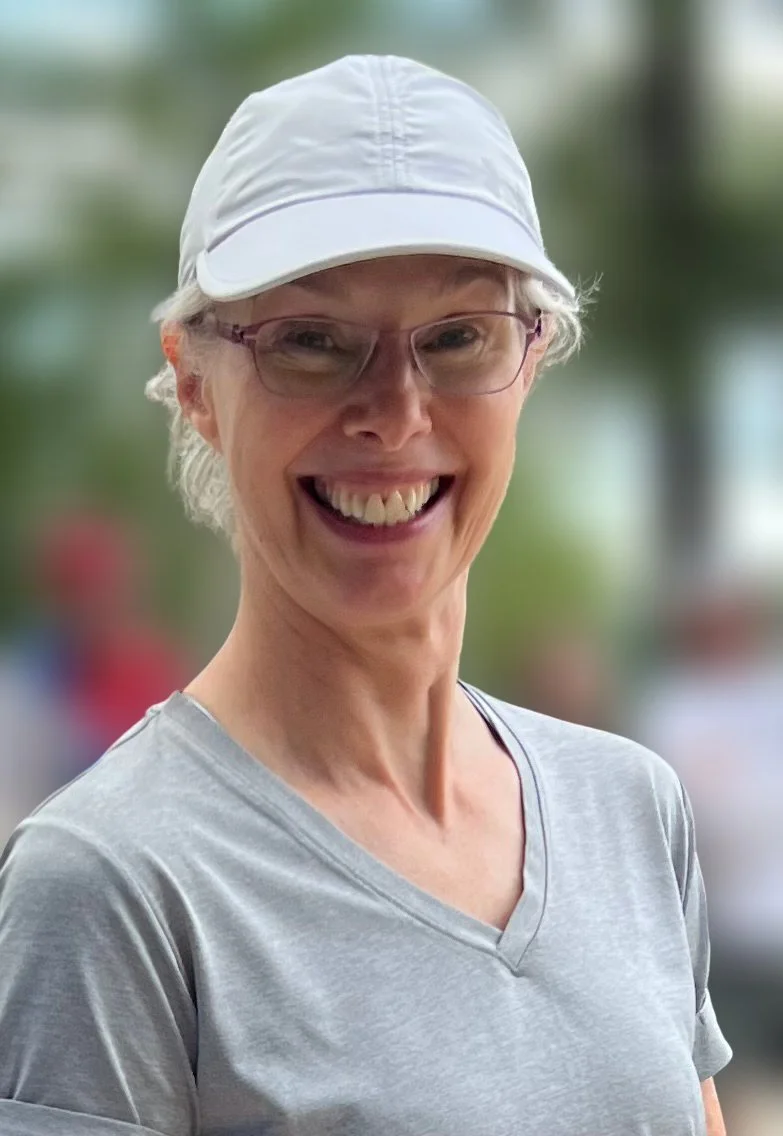 A smiling older woman wearing glasses, a gray T-shirt, and a white cap outdoors with blurred background.