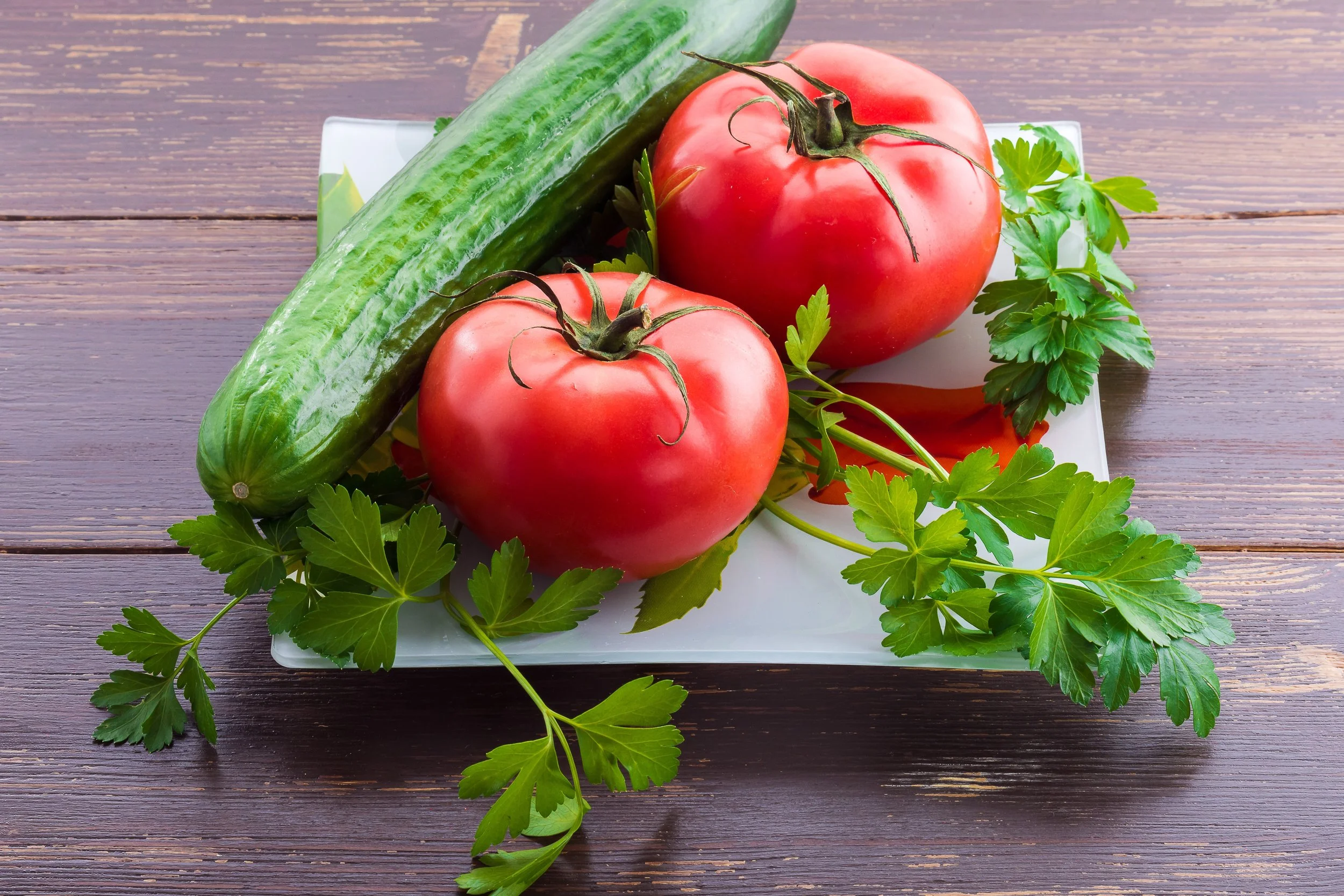Fresh cucumber, two ripe tomatoes, and sprigs of parsley on a white plate on a wooden surface.
