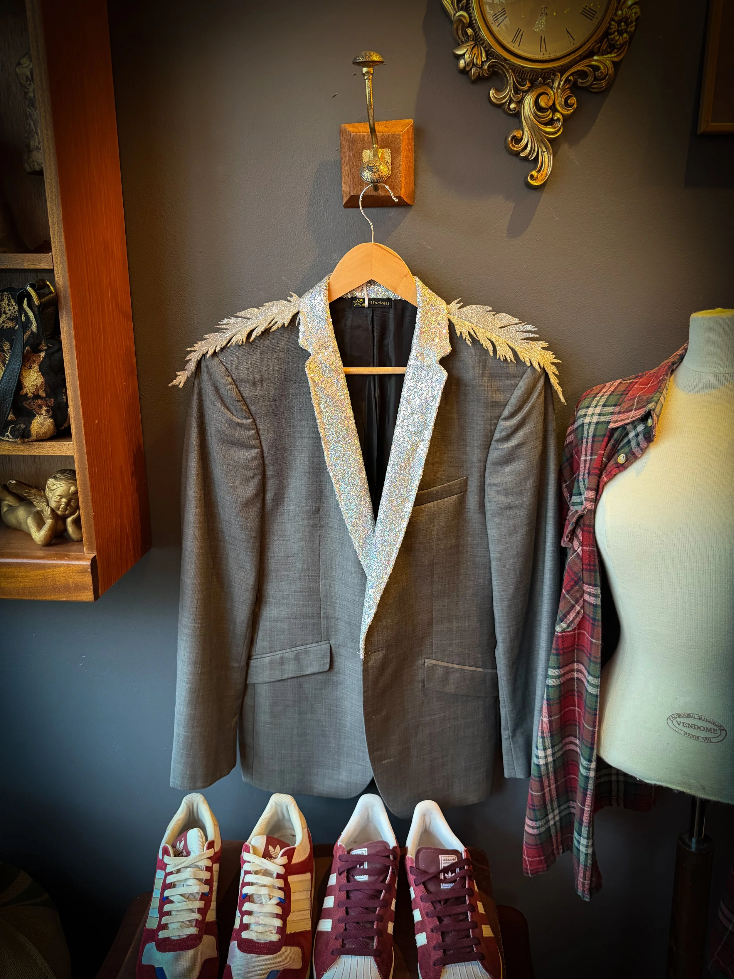 A mannequin wearing a plaid shirt next to a brown blazer with glittery lapel and shoulder details, hanging on a hanger on a wall. A pair of Adidas sneakers with red stripes and white laces are on a table below.