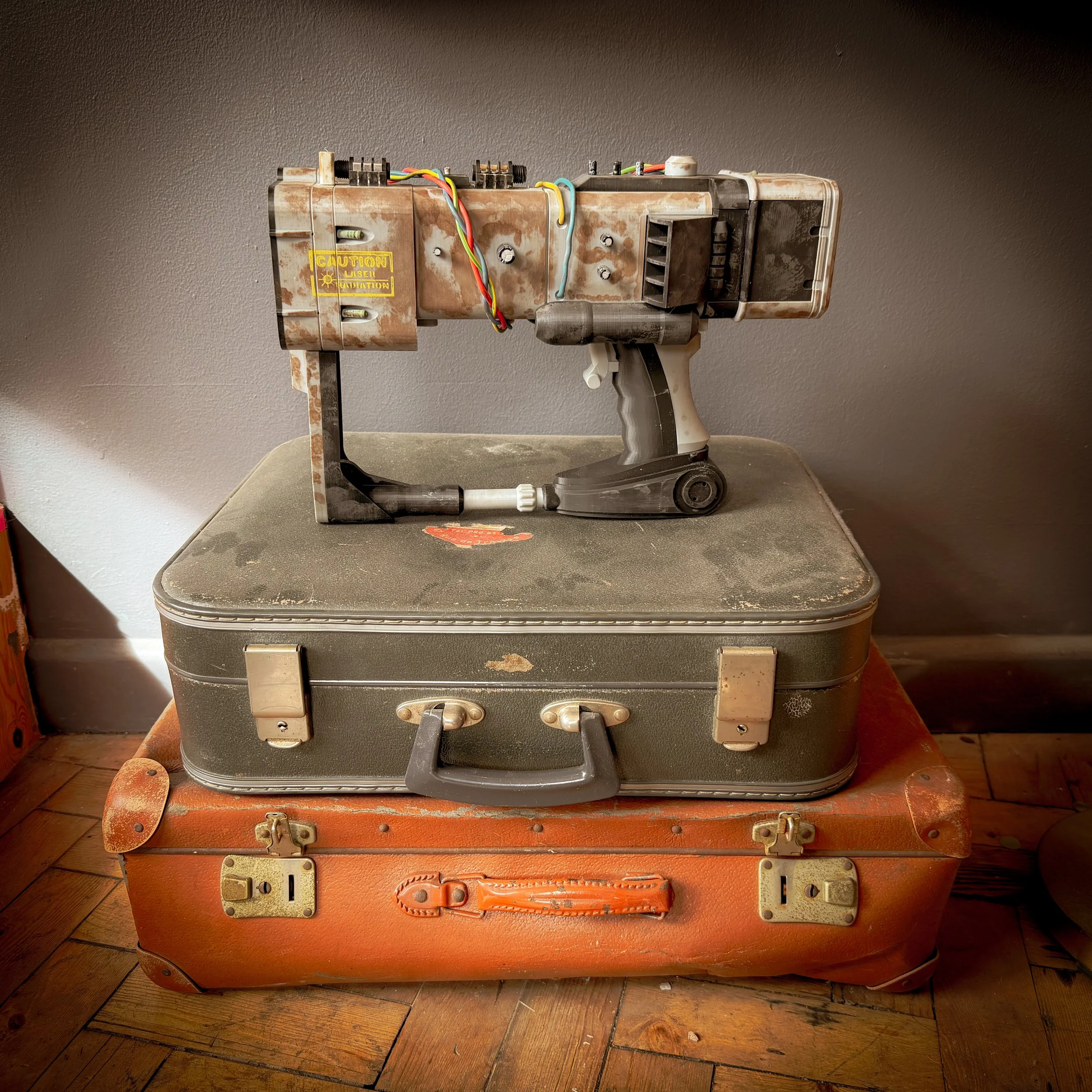 A vintage laser gun prop mounted on top of two stacked old suitcases, one greenish gray and one orange, sitting on a wooden floor against a gray wall.