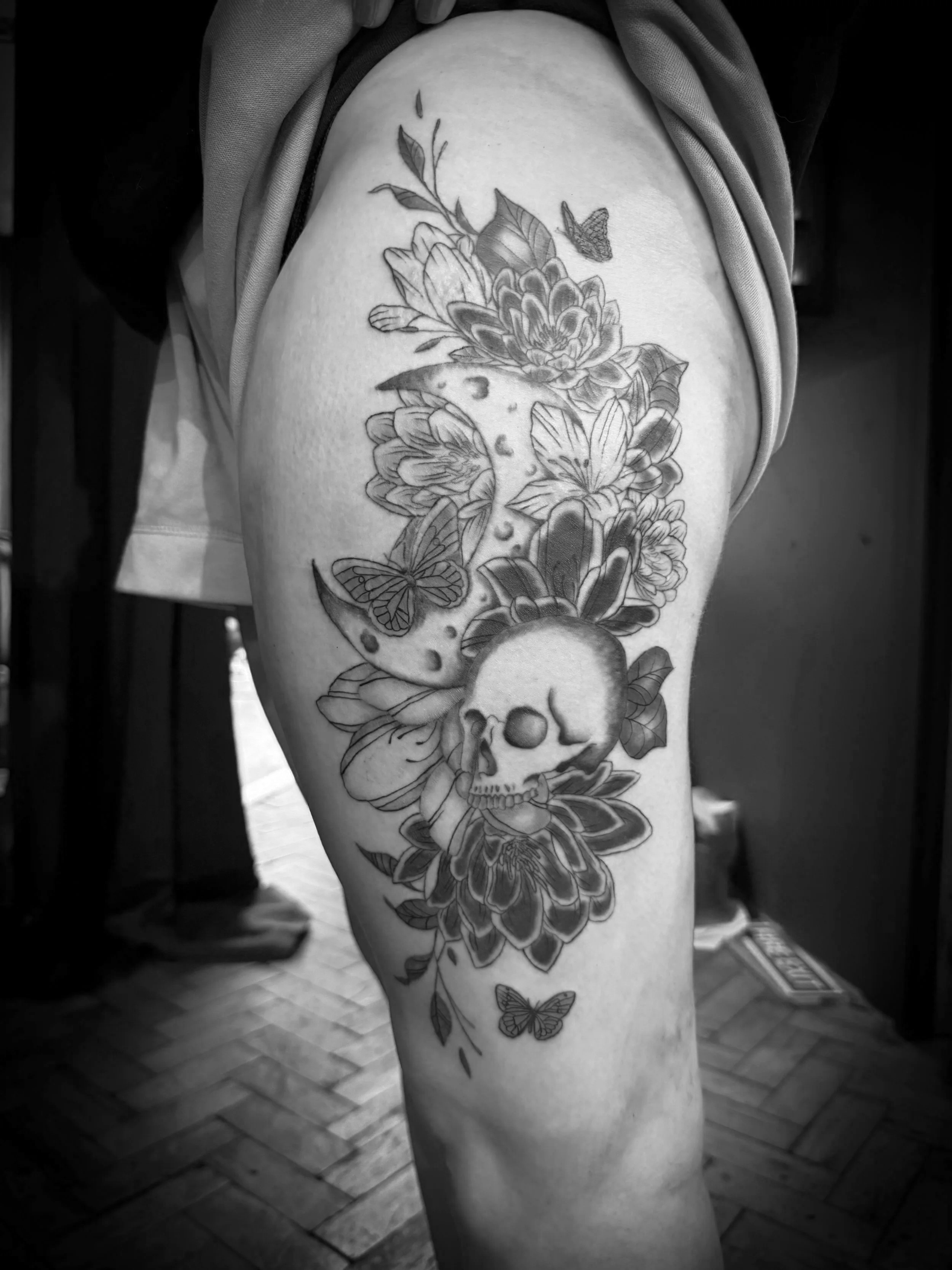 A person's upper thigh and hip with a large tattoo featuring flowers, butterflies, and a skull, in black and white.