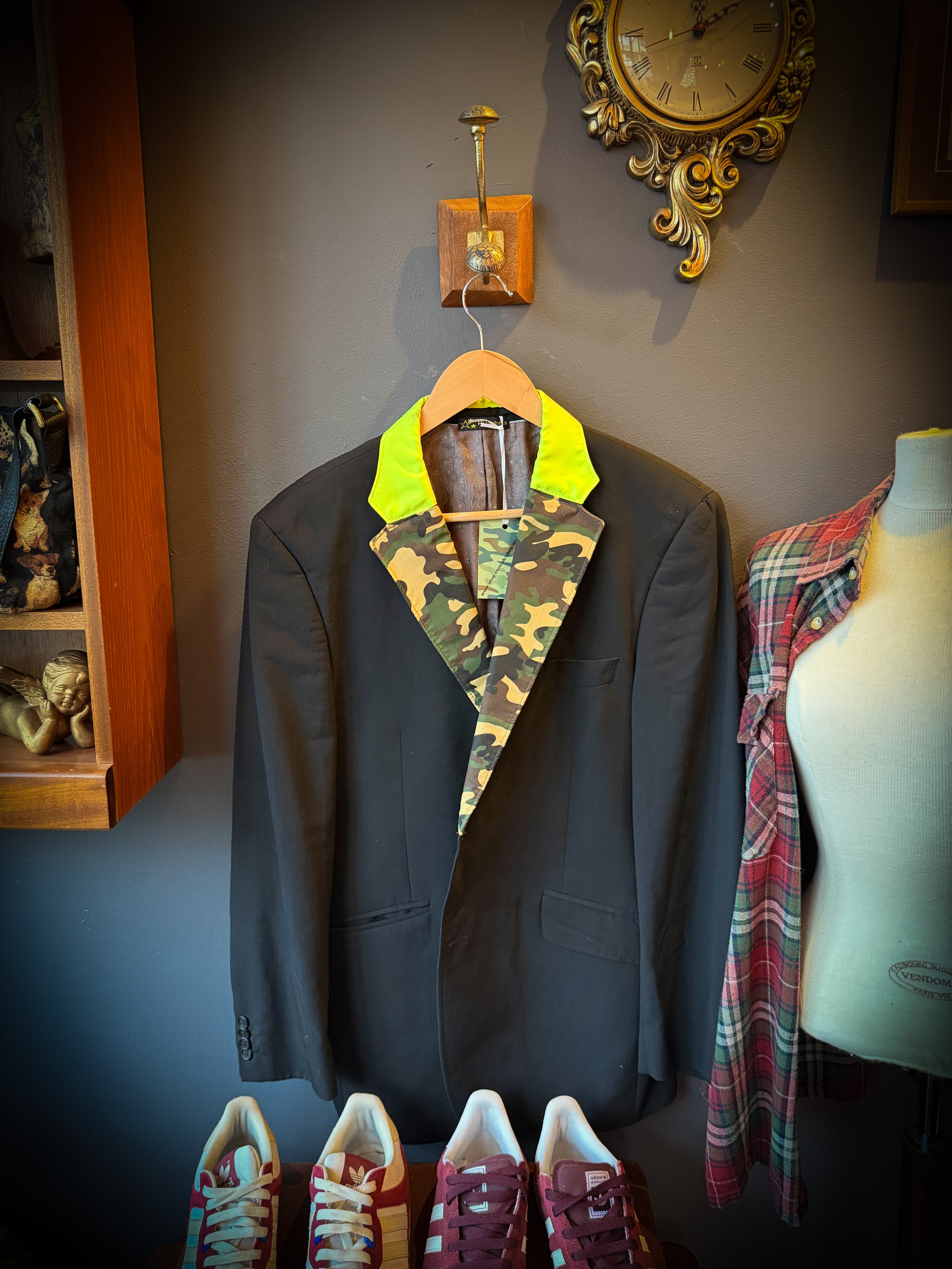 A black blazer with a camouflage collar hanging on a wooden hanger, displayed against a gray wall. A mannequin with a plaid shirt is partially visible on the right. A pair of red and white sneakers is at the bottom of the image, and a vintage clock i