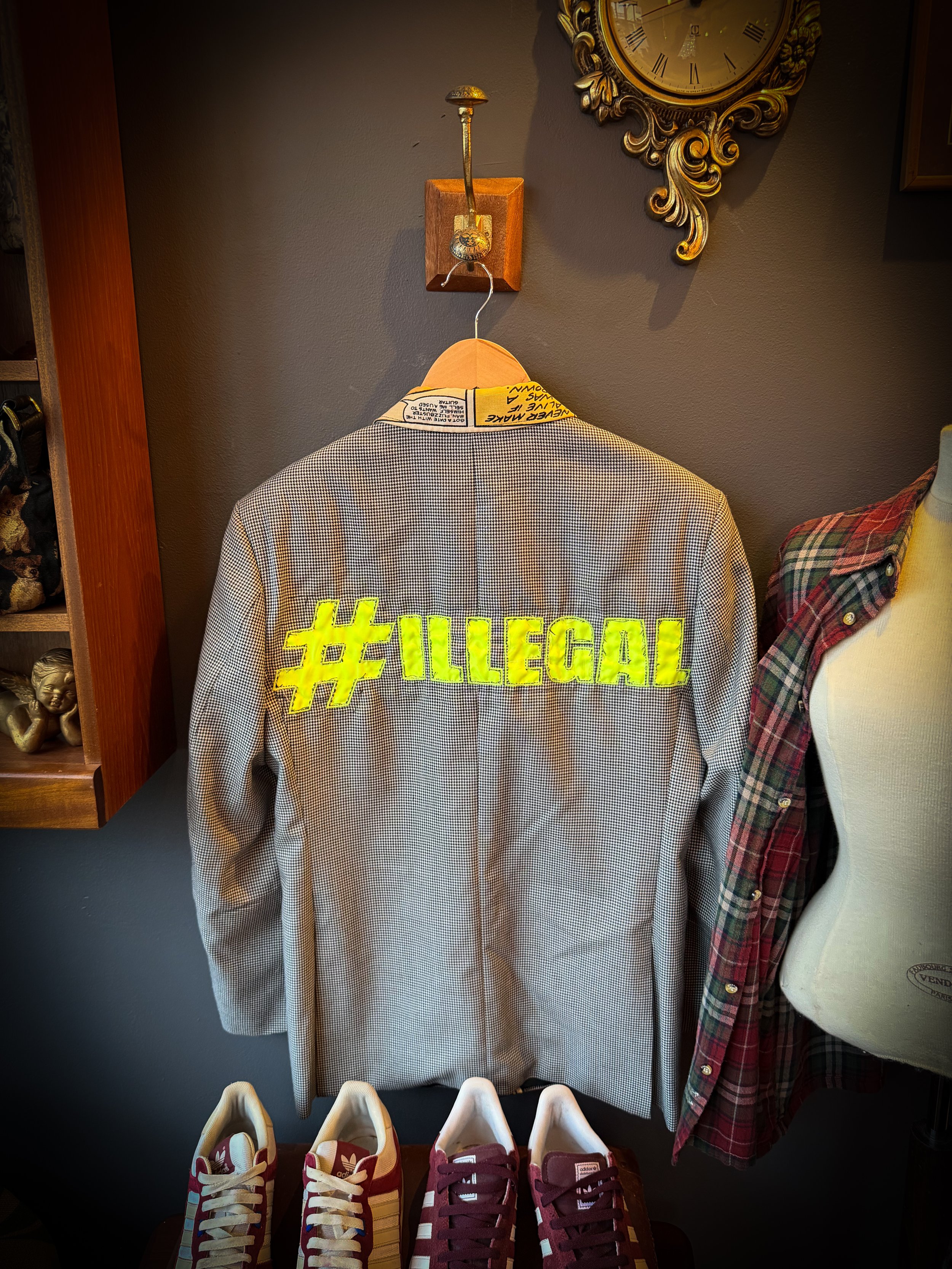 A gray blazer with bright yellow letters that read '#ILLEGAL' on the back, hanging on a hanger against a gray wall. Shoes are placed below it, and part of a plaid shirt and mannequin are visible on the right. A vintage clock is on the wall above.