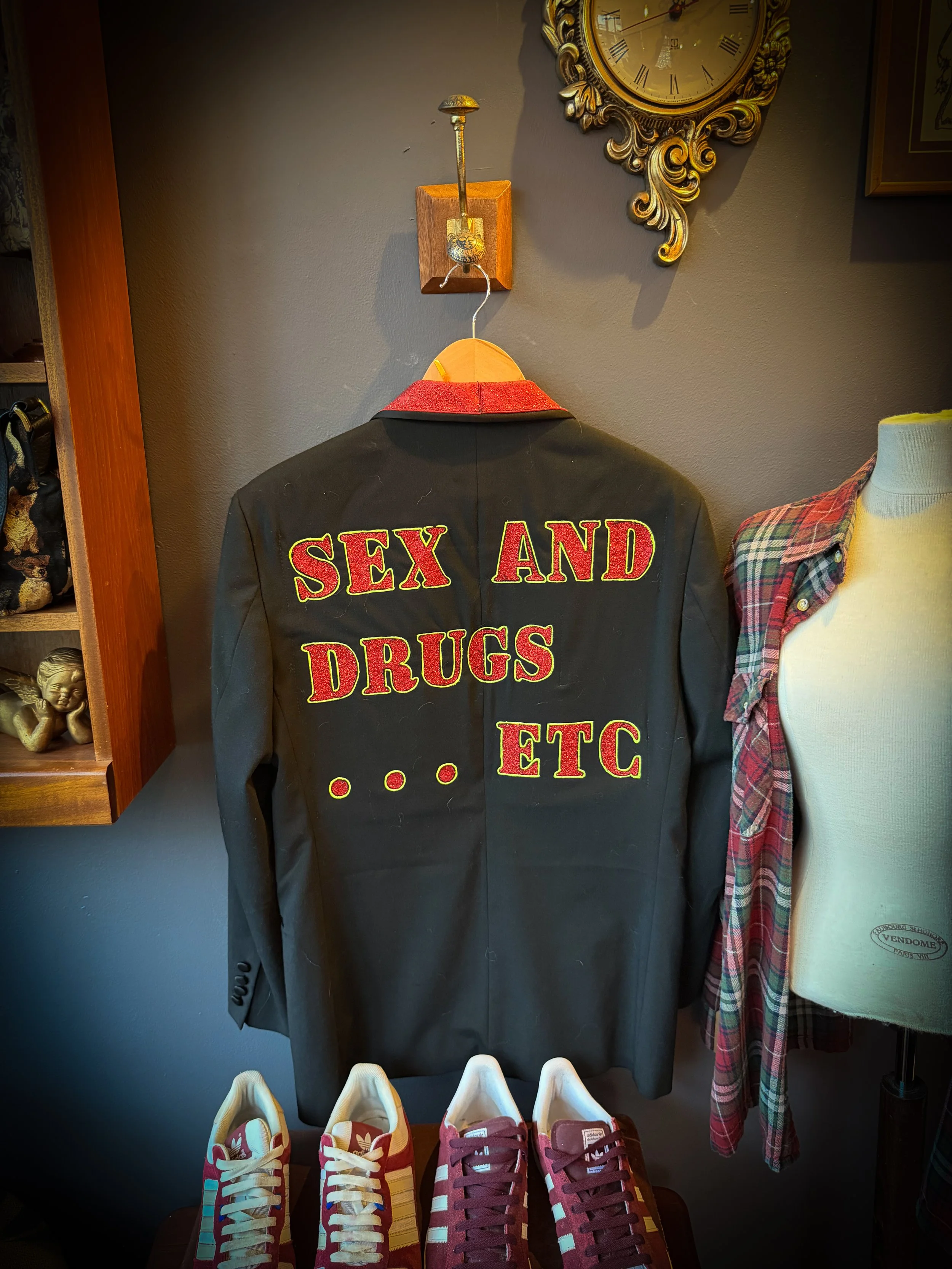 Back of a black jacket with yellow and red embroidered text reading "SEX AND DRUGS ETC," hanging on a wooden hanger on the wall. There are sneakers at the bottom and a mannequin torso to the right.
