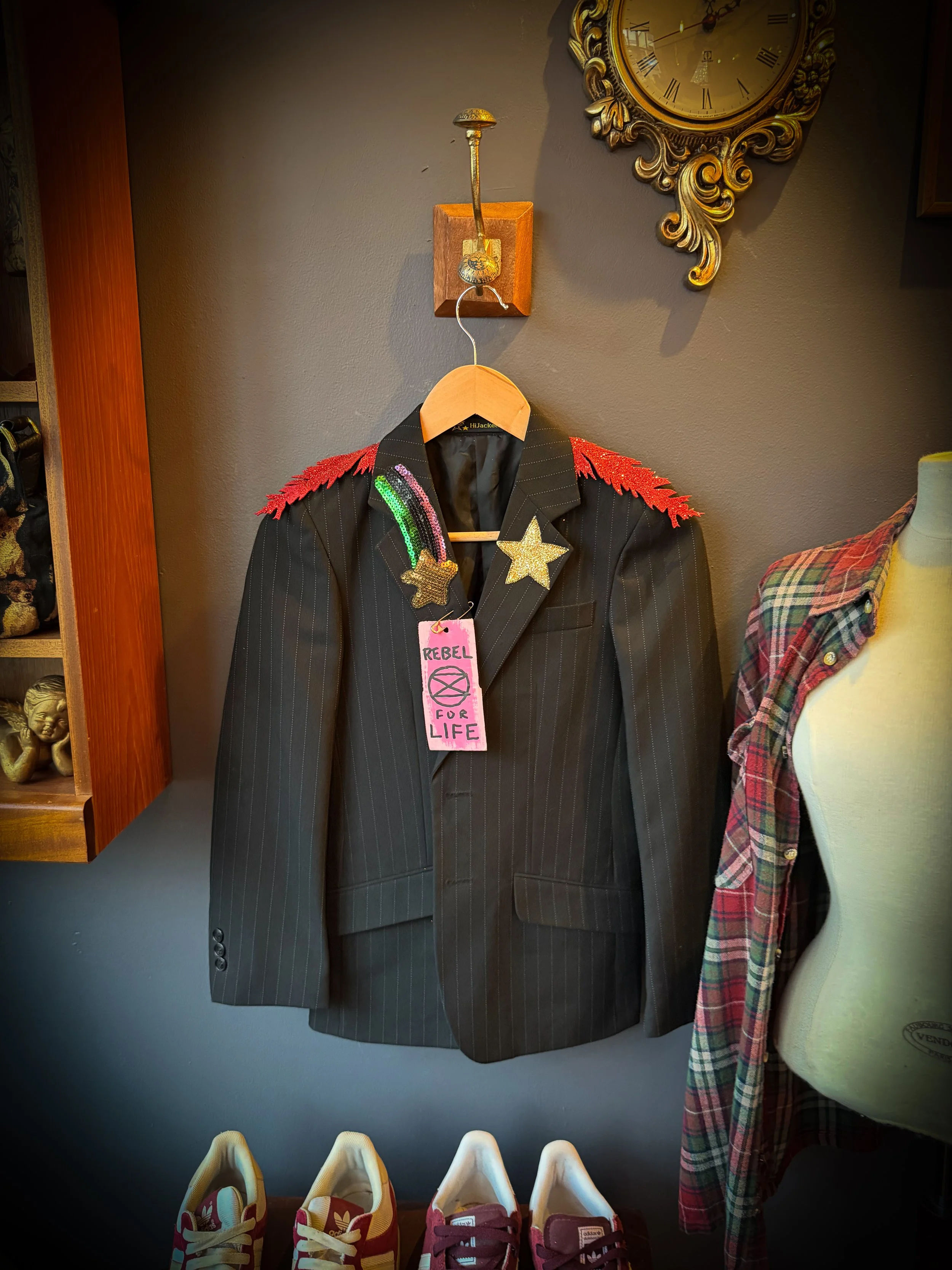A pinstripe blazer decorated with patches like a rainbow, stars, and red shoulder accents, hanging on a wall with a pink tag reading 'Rebel for Life', below a vintage clock and an ornate wall clock, with sneakers on the floor.