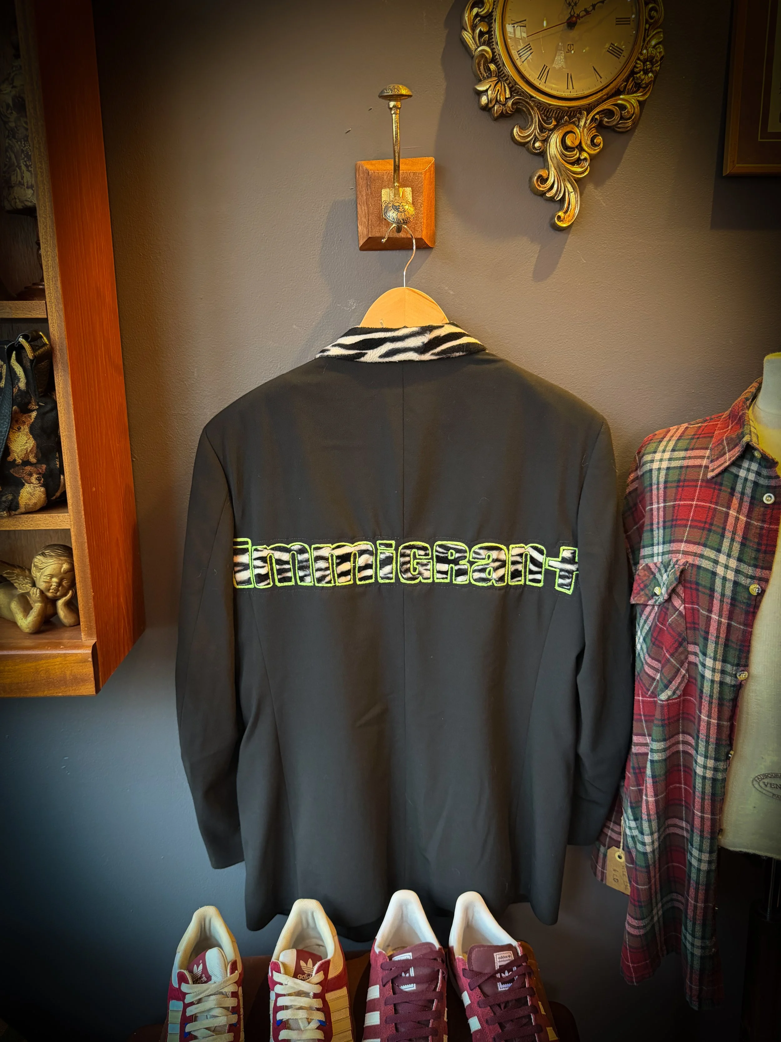 A black jacket with a zebra-striped collar hanging on a wall with the word 'immigrant' embroidered on it. There are red and white sneakers in front of the jacket and a plaid shirt on a mannequin to the right.
