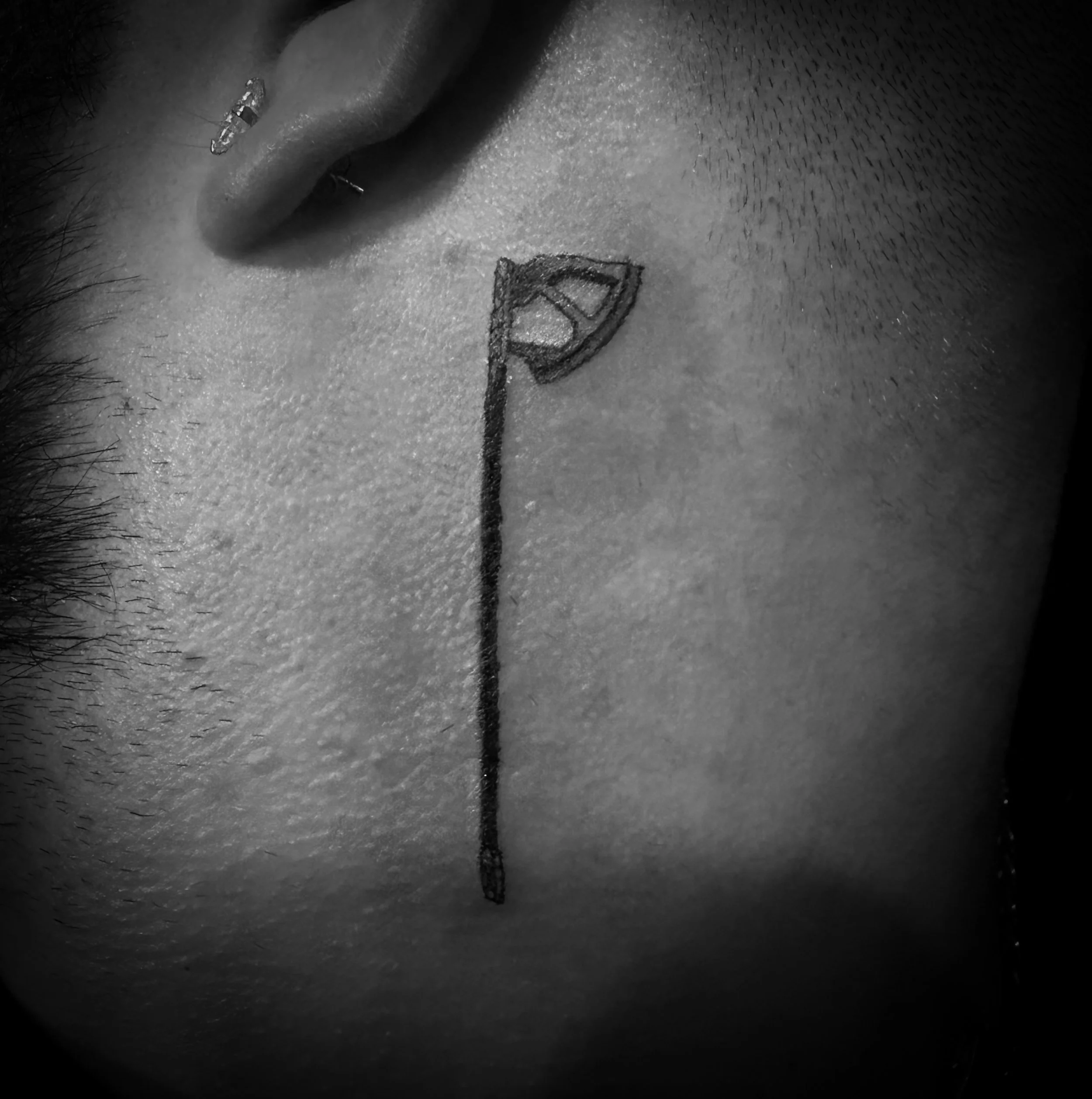 A close-up black and white photo of a person's face featuring a small flag tattooed on their cheek, a piercing in the ear, and short facial hair.