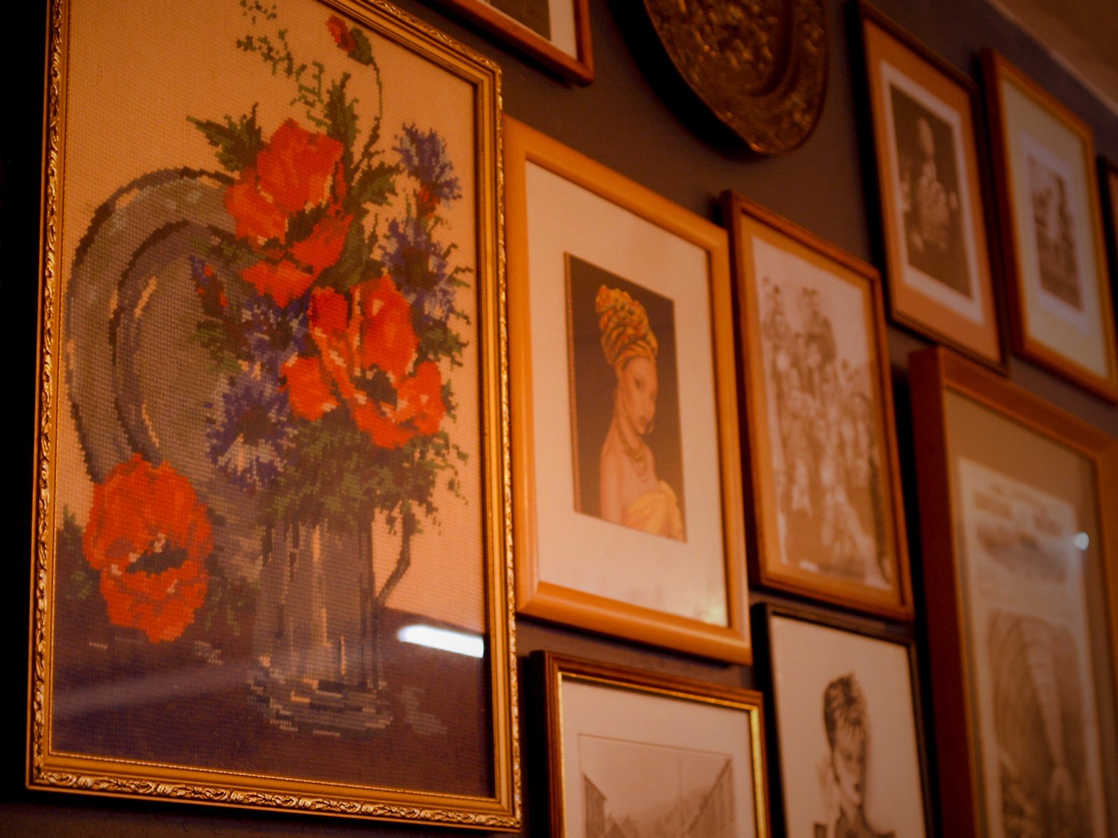 A gallery wall with multiple framed artworks including a floral cross-stitch picture, a portrait of a woman with a headwrap, and various other paintings and photographs in gold and wooden frames.