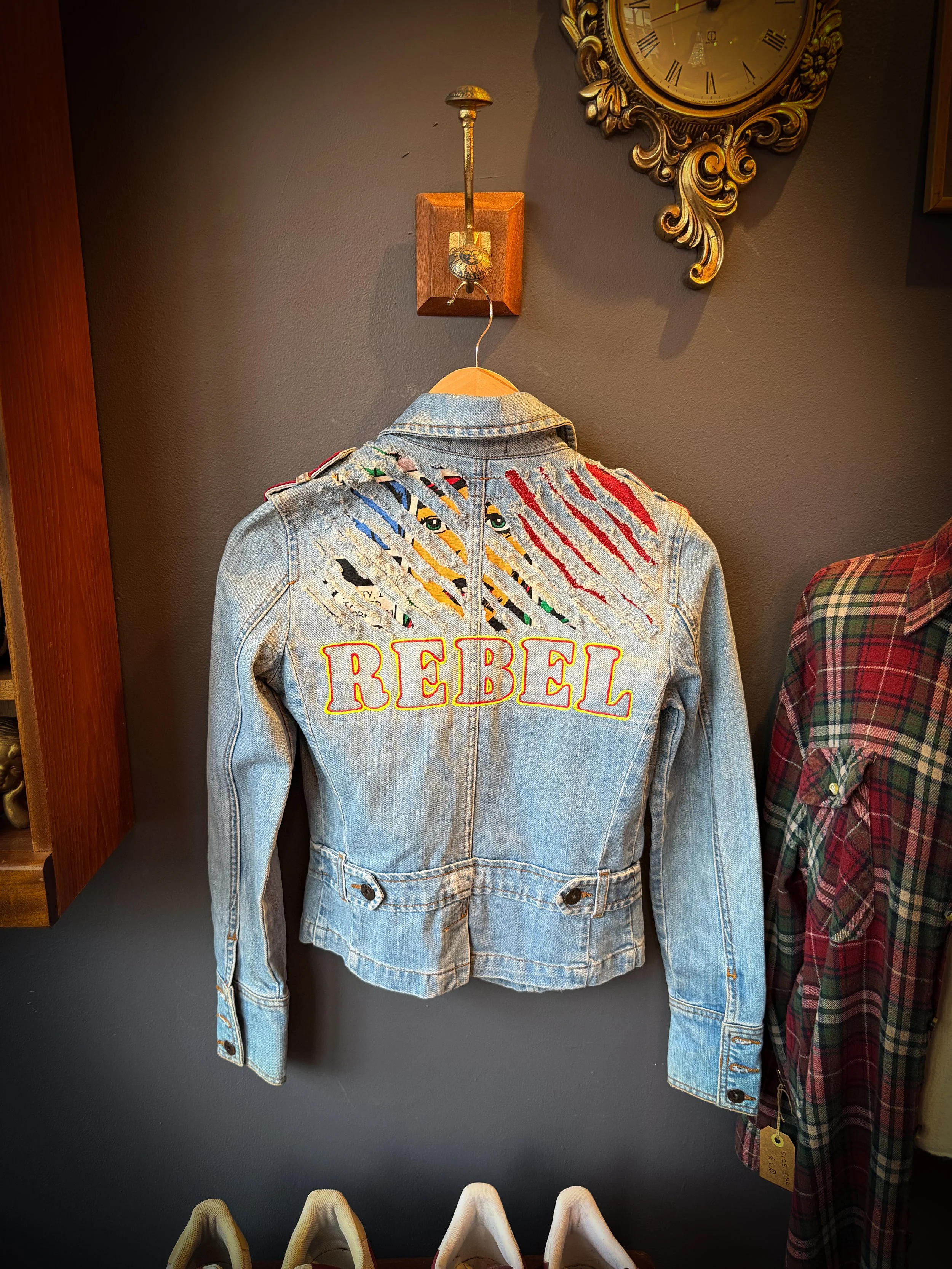 Denim jacket with the word 'REBEL' embroidered on the back, hanging on a wall in a store.