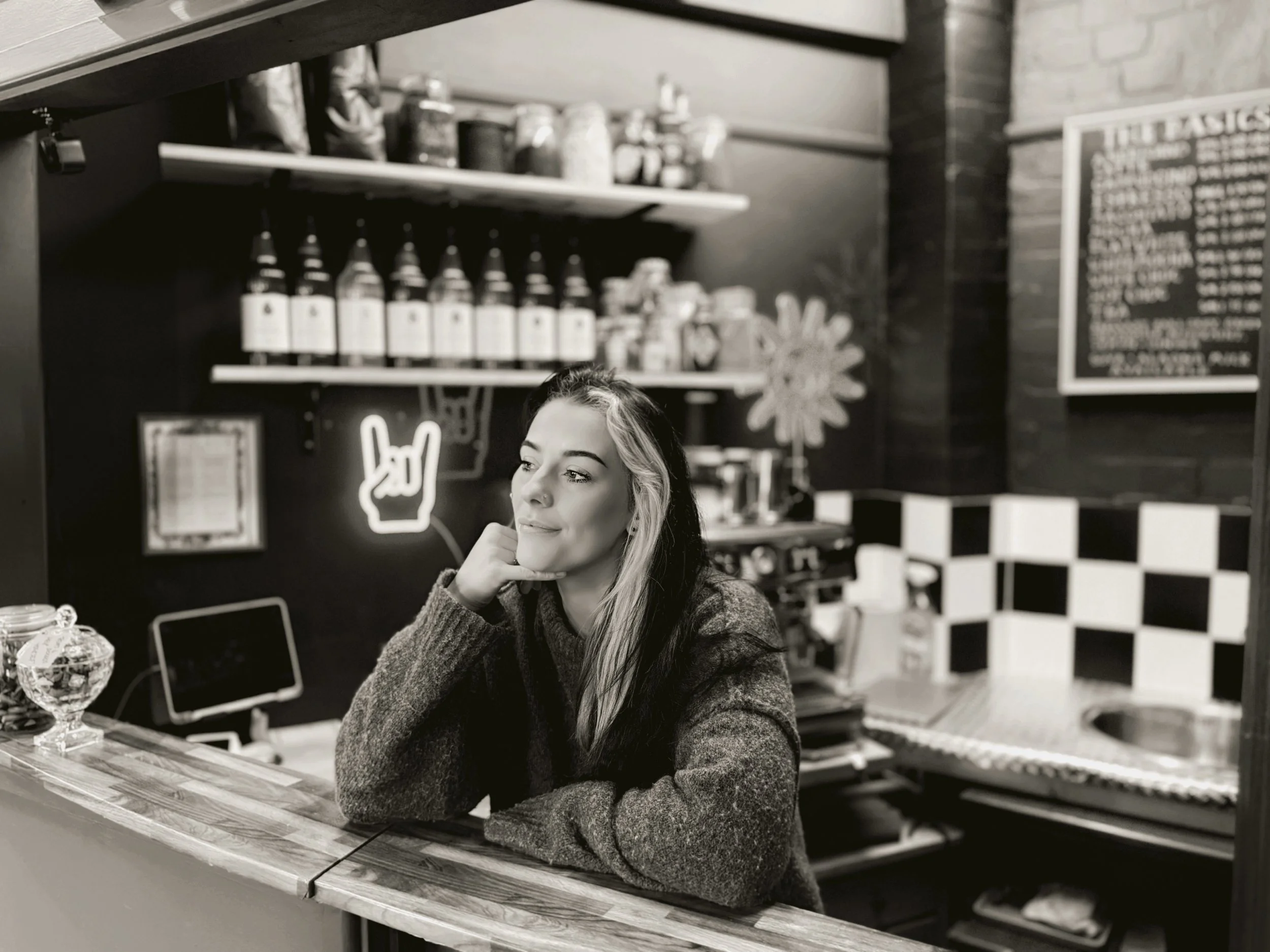 A woman with black and blonde hair sitting at a bar counter, resting her chin on her hand, in a cafe or bar setting, with bottles on a shelf and a menu on the wall behind her.