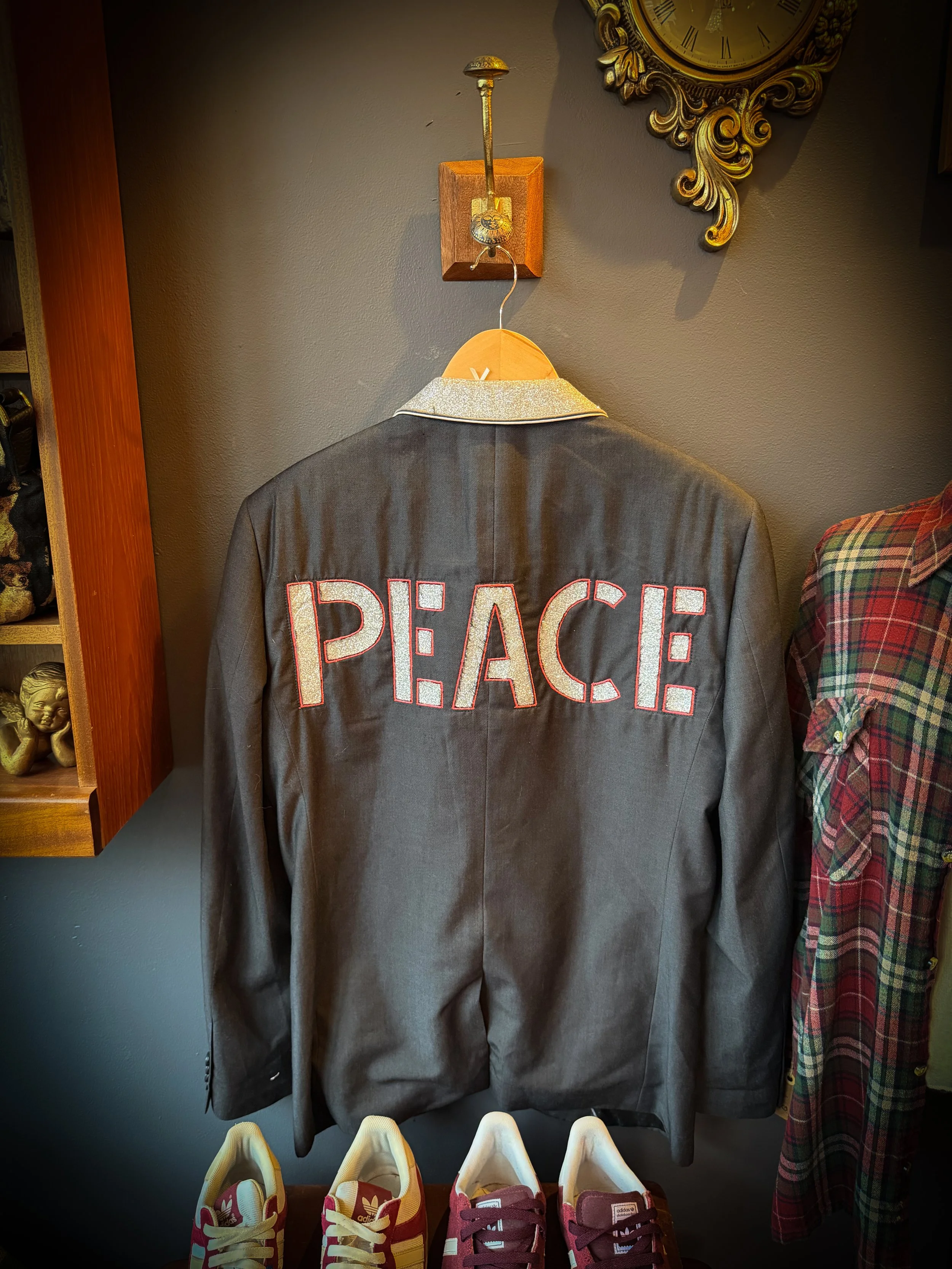 Back of a dark gray blazer with the word 'PEACE' spelled out in large, glittery letters on the back, hanging on a wooden hanger on a hook on a wall, with red and yellow sneakers below and a plaid shirt to the right, with a decorative gold clock above