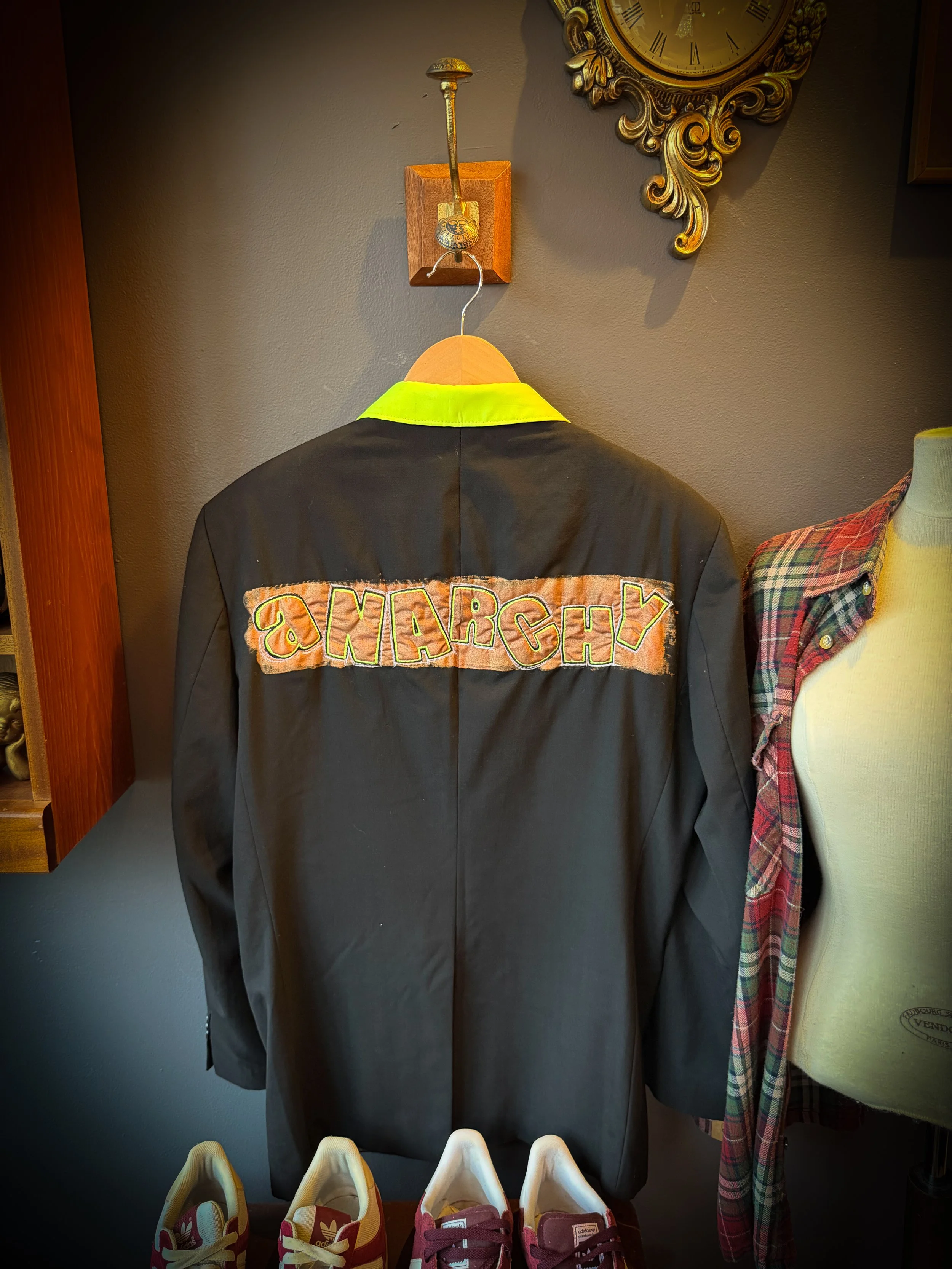 Black jacket with orange and green 'MARCH' patch on back, hanging on a wooden hanger against a gray wall, with a mannequin wearing a plaid shirt and red sneakers in front.