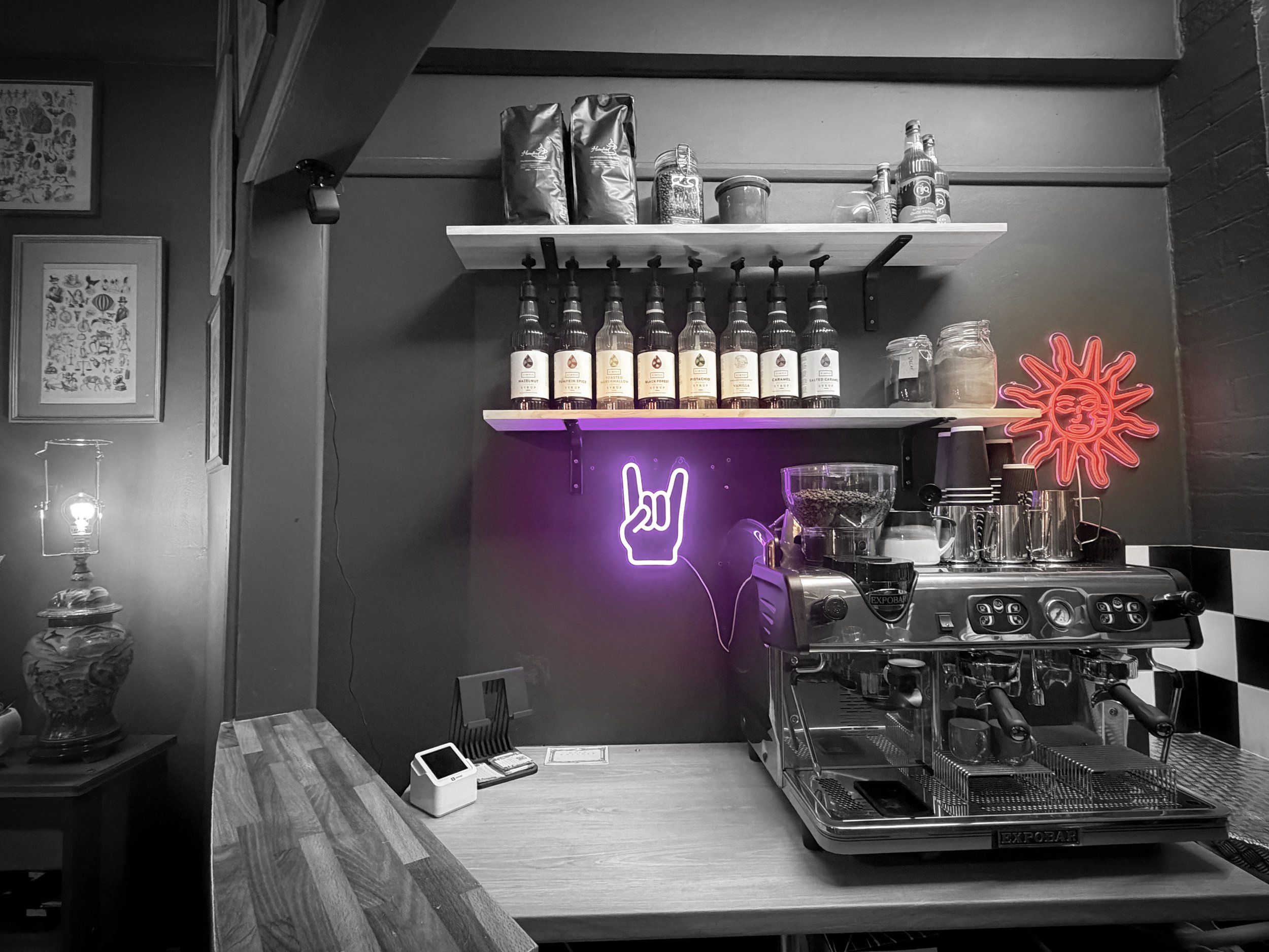 Coffee shop counter with an espresso machine, a neon sun sign, and a neon hand sign in the shape of a rock and roll gesture.