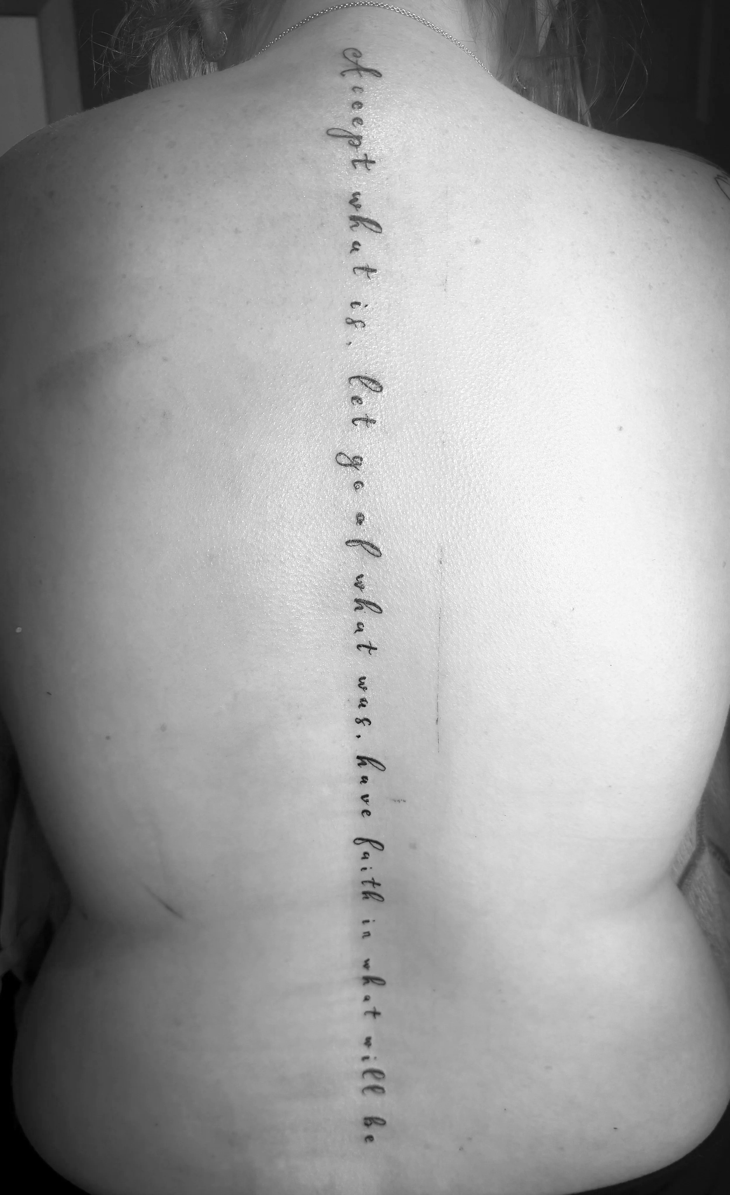 Back of a person's upper back with a vertical tattoo of cursive handwriting that reads 'If I got it, what if I do? I want what you can give.'