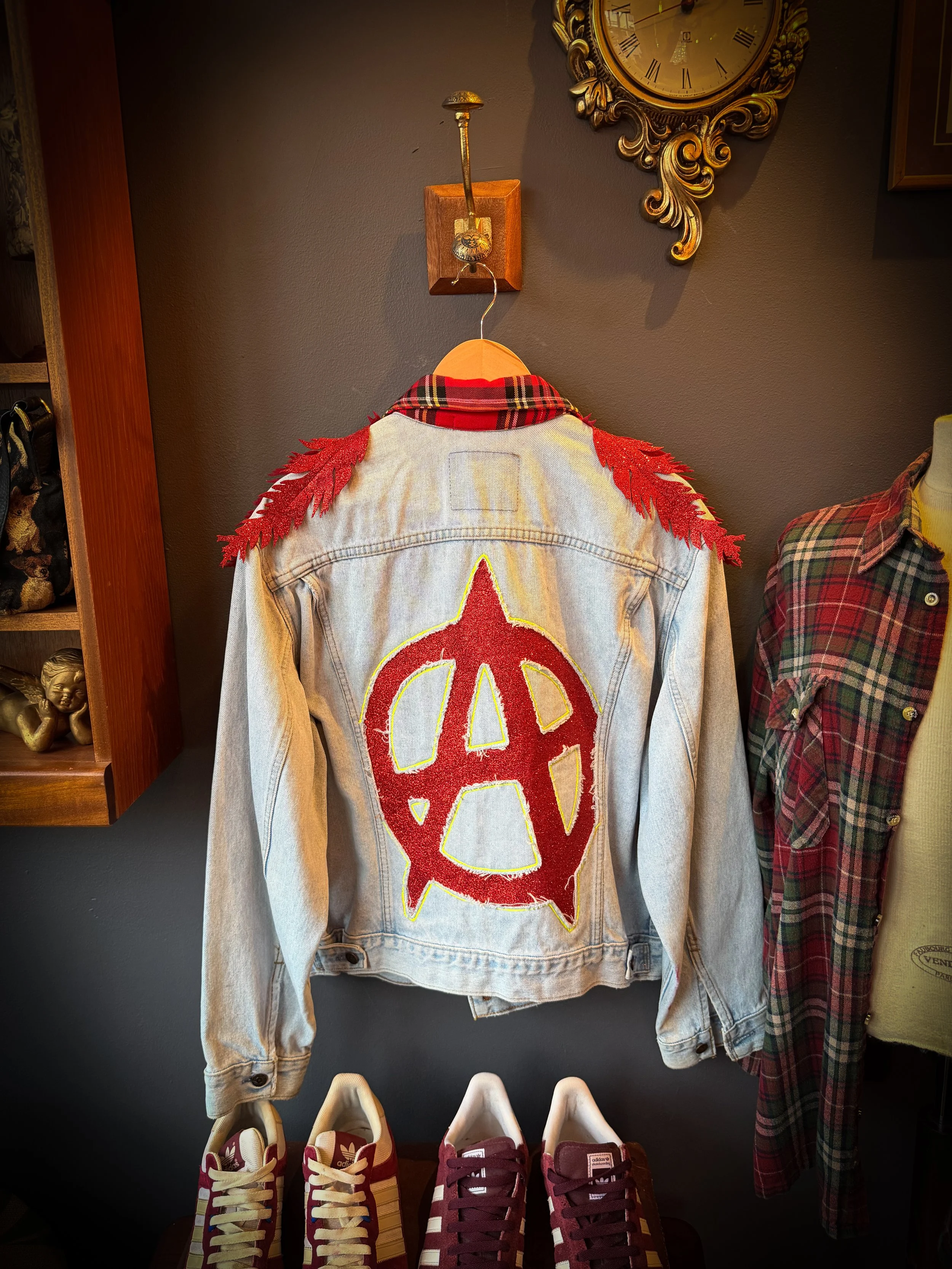 A denim jacket with red fringe shoulder accents and a large red and yellow anarchist symbol patch on the back, hanging on a wooden hanger on a wall with a decorative clock above.
