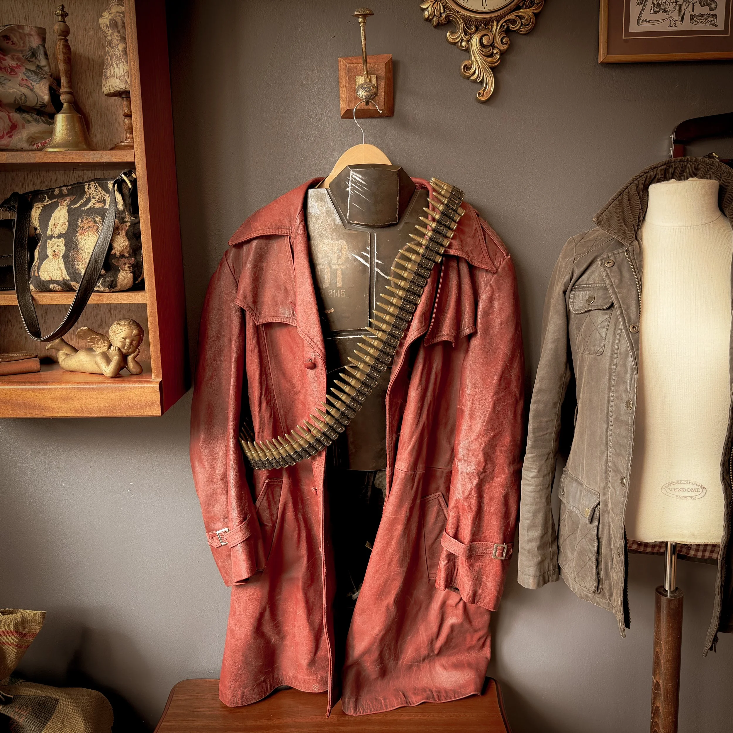 A vintage military display including a mannequin with a red leather trench coat and bullet belt, and a gray jacket on a separate mannequin, with a wall clock and shelves with various items in the background.