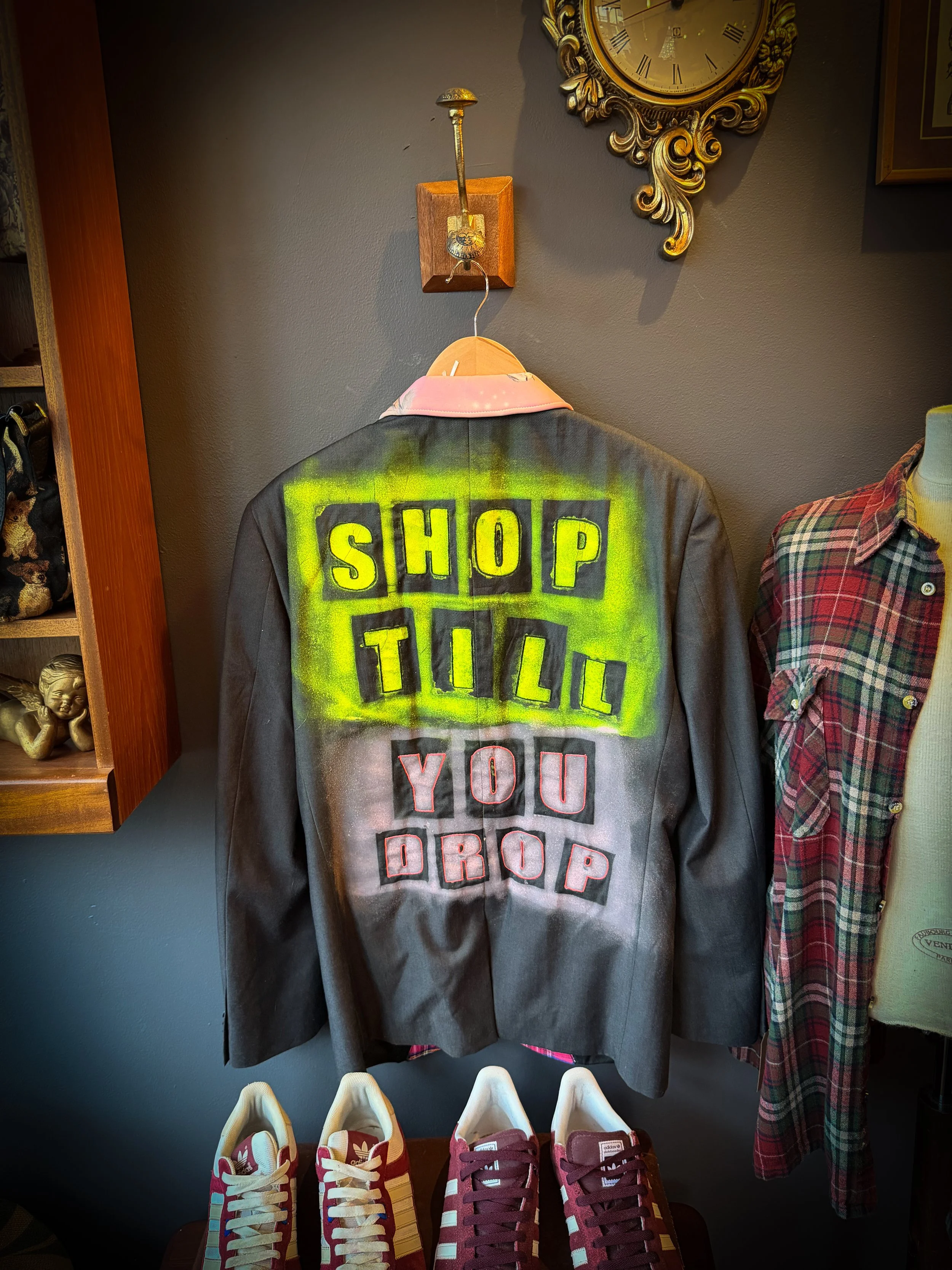 A vintage leather jacket with graffiti-style painted words that say "SHOP TILL YOU DROP" on the back, displayed on a hanger. The jacket is matching with a pink collared shirt underneath, and there are red and white sneakers placed below the jacket.