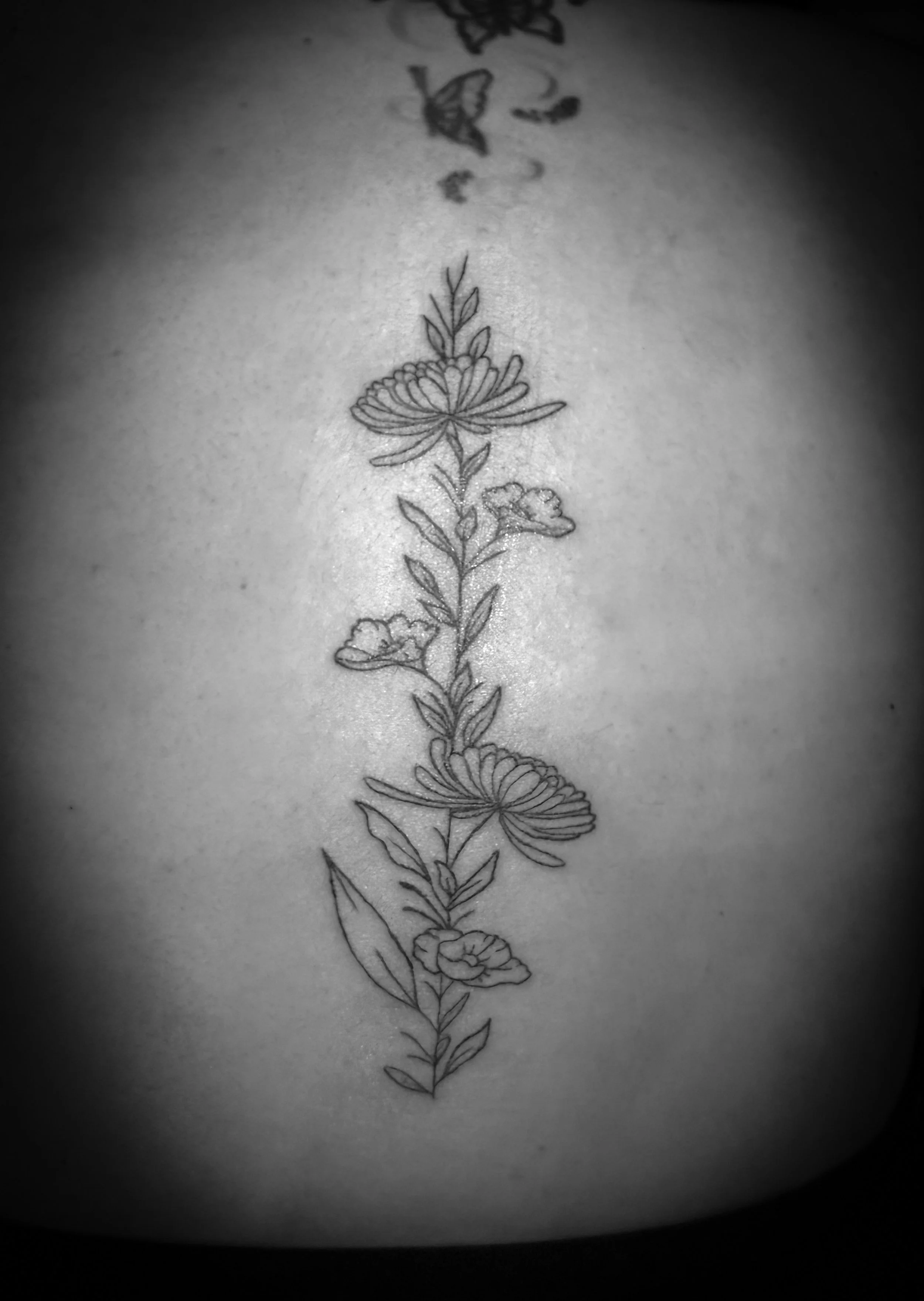 Black and white photo of a delicate floral tattoo on skin, featuring various flowers and leaves arranged vertically.
