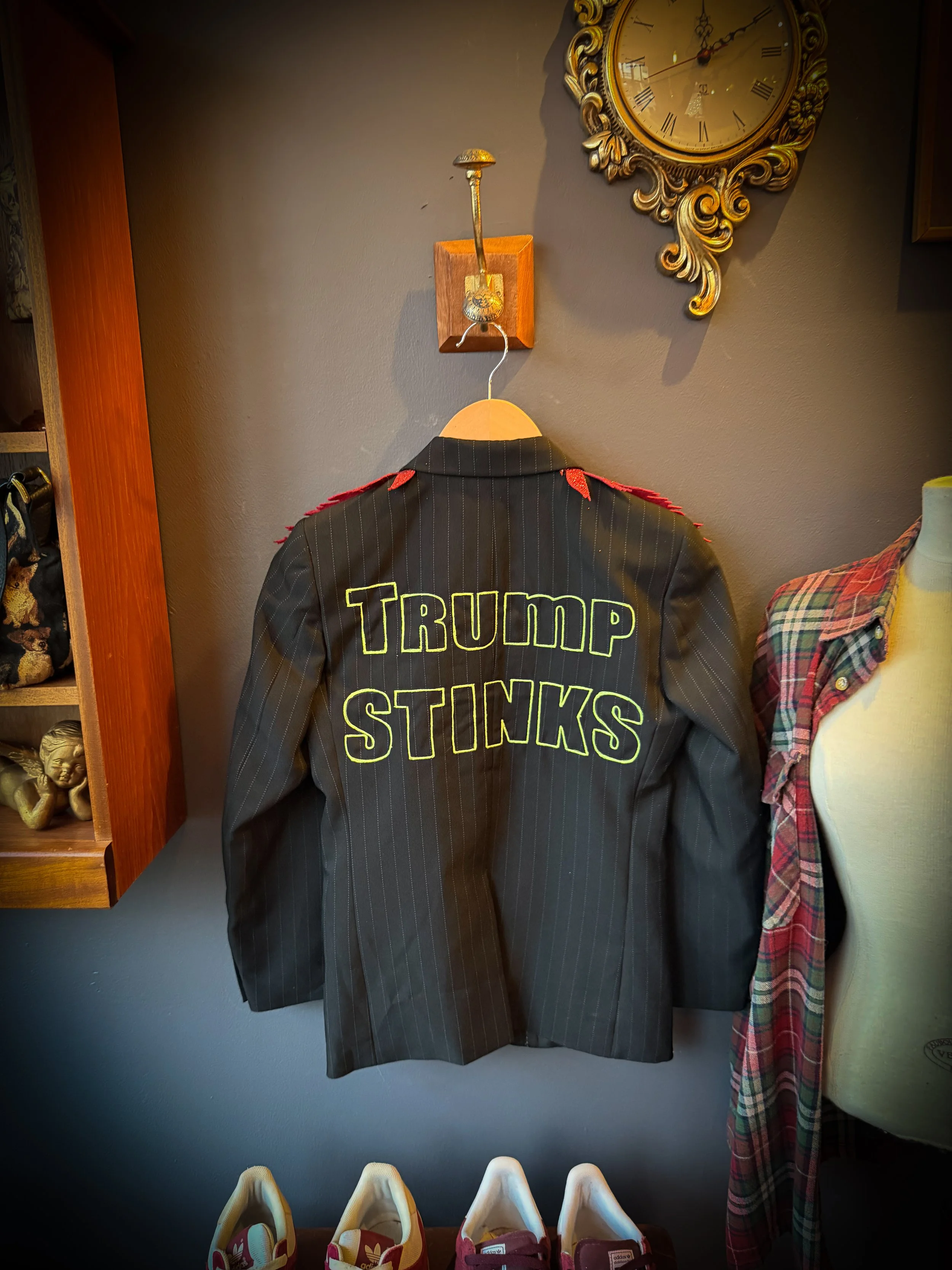 A jacket hanging on a wall with the words 'TRUMP STINKS' embroidered on the back in large yellow and black letters, with red epaulettes on the shoulders. The image also shows parts of a clock, a mannequin with a plaid scarf, and shelves with decorati