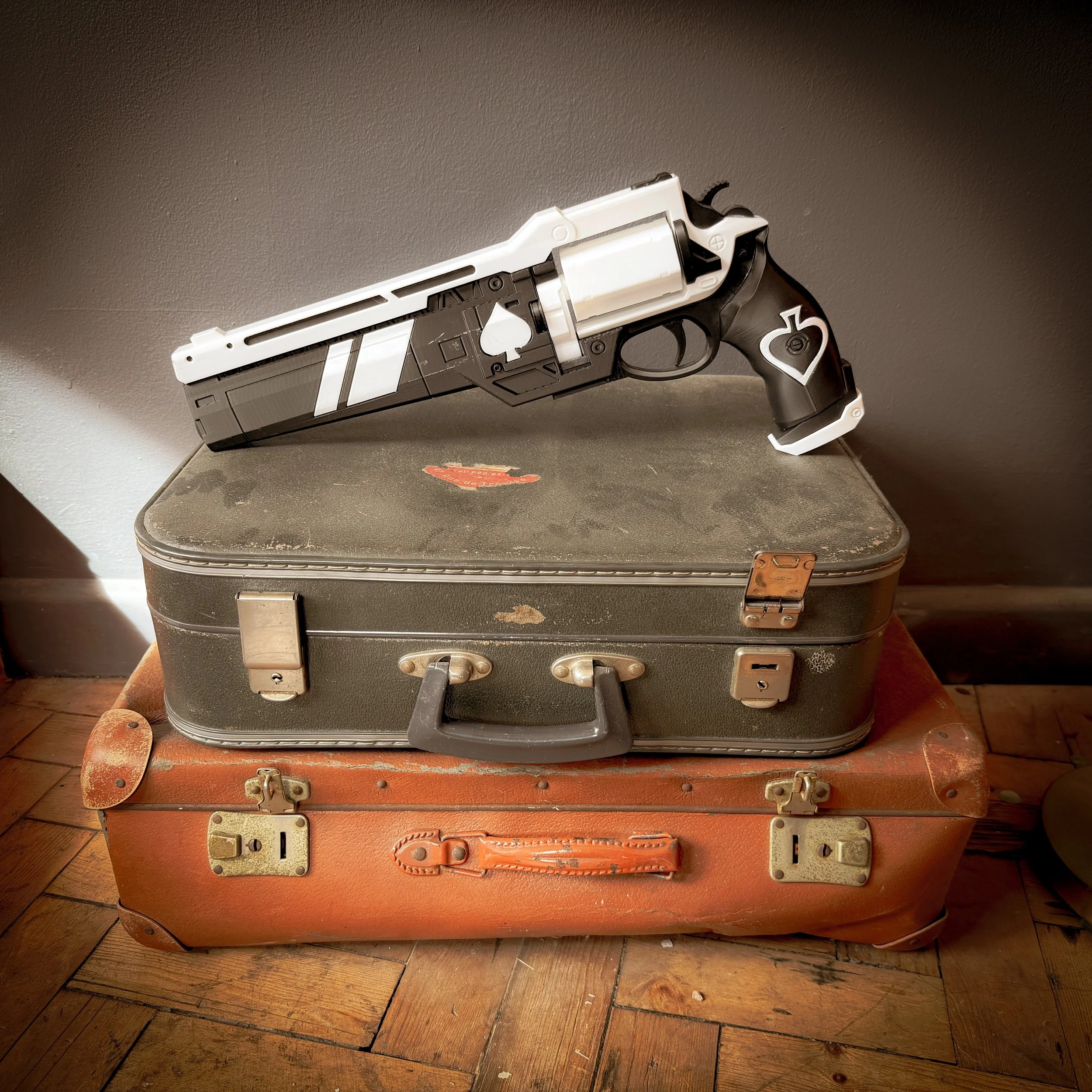 A black and white revolver with playing card symbols rests on top of two vintage suitcases stacked on a wooden floor.