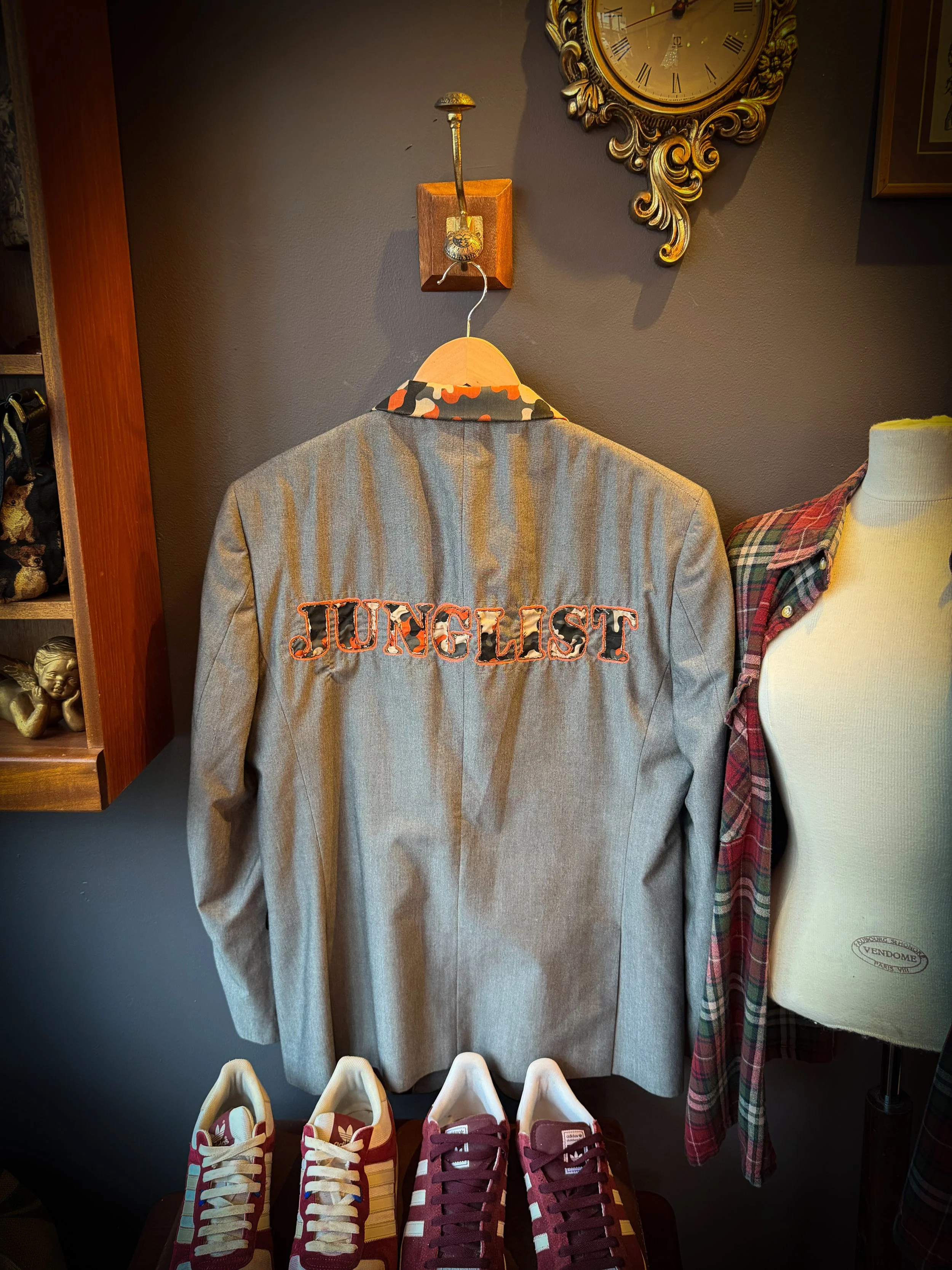 A beige blazer with 'JUNGLEST' embroidered on the back hangs on a hanger against a dark wall, with a clock above it and sneakers below.