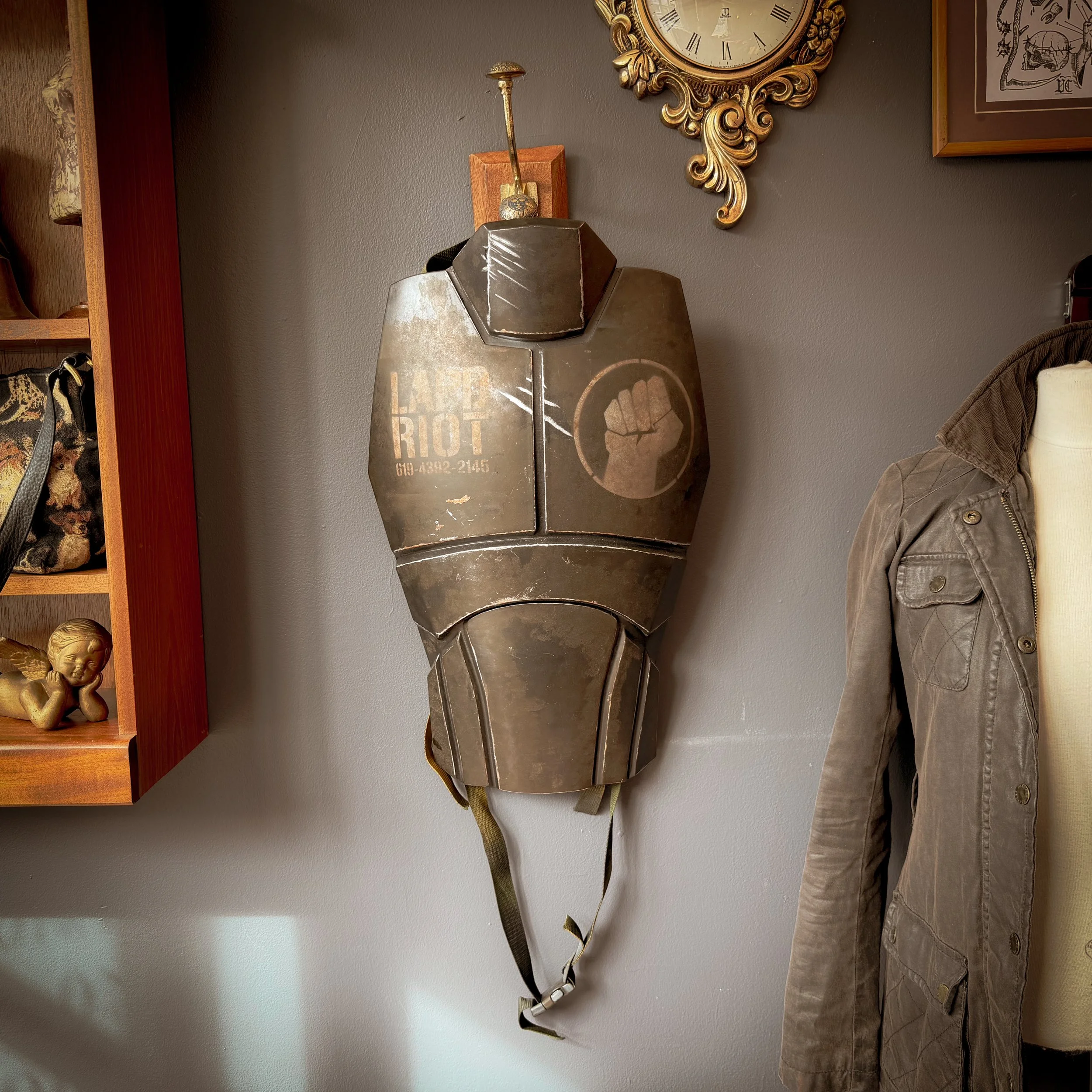 A vintage tactical vest with a fist symbol and the word 'Riot' written on it, hanging on a gray wall.