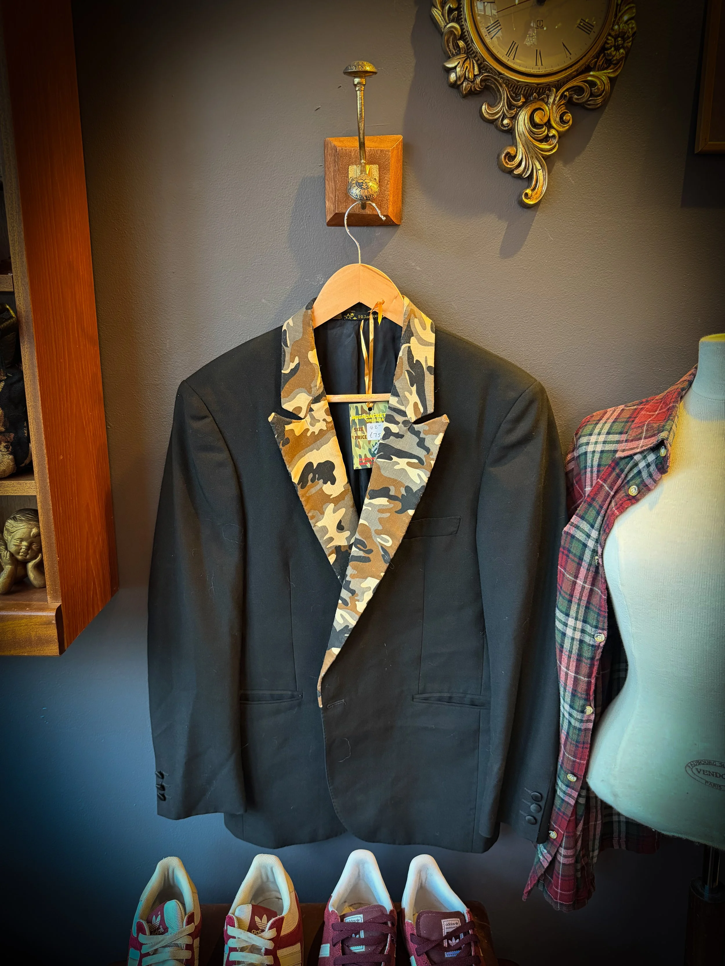 Black blazer with camouflage pattern lapel hanging on a wooden hanger, displayed on a wall in a store.