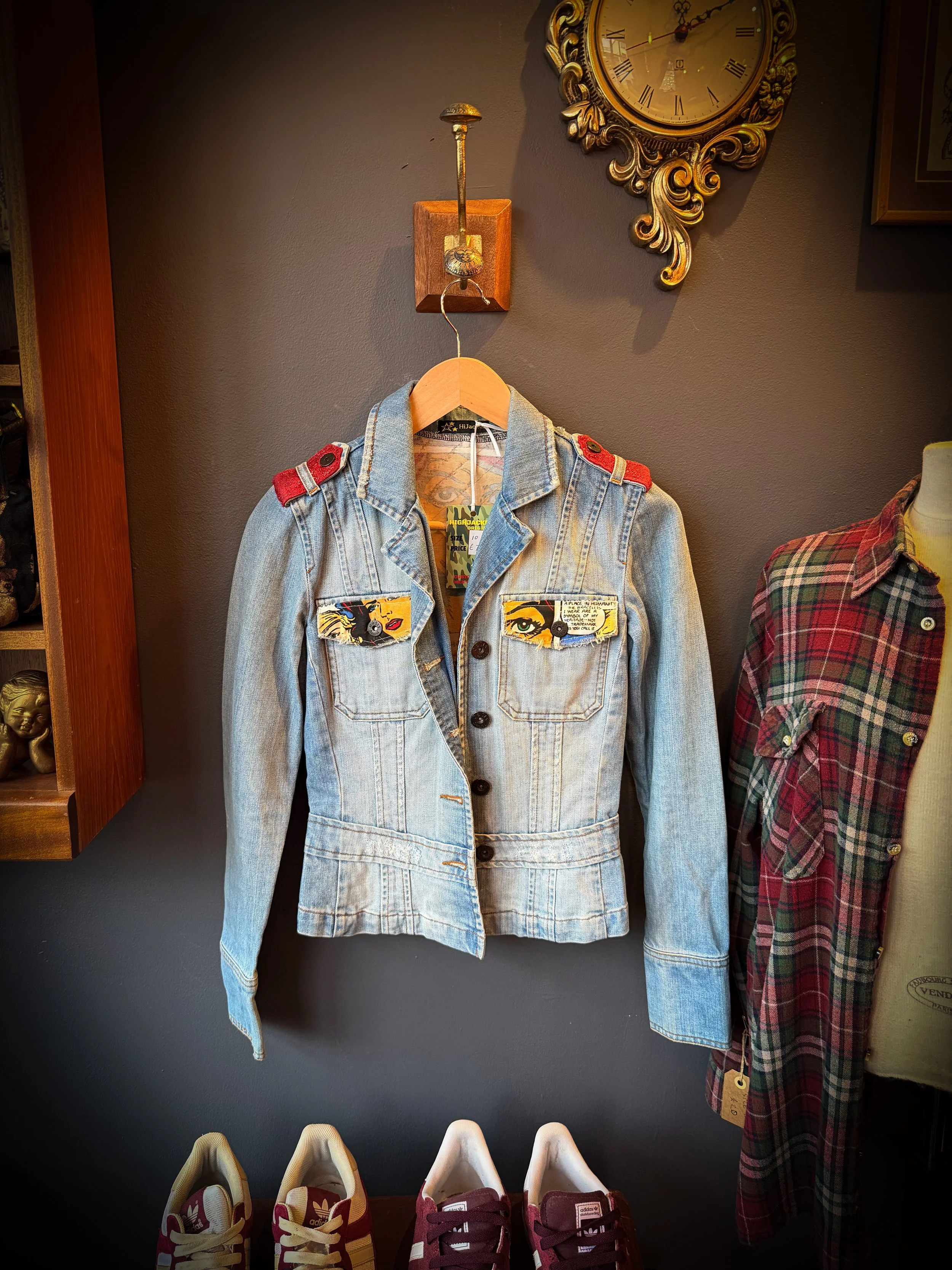 A denim jacket with comic book style patches on the front pockets, hanging on a wooden hanger. The jacket has red shoulder epaulets. There are vintage shoes at the bottom, and a wall clock above with a gold ornate frame.