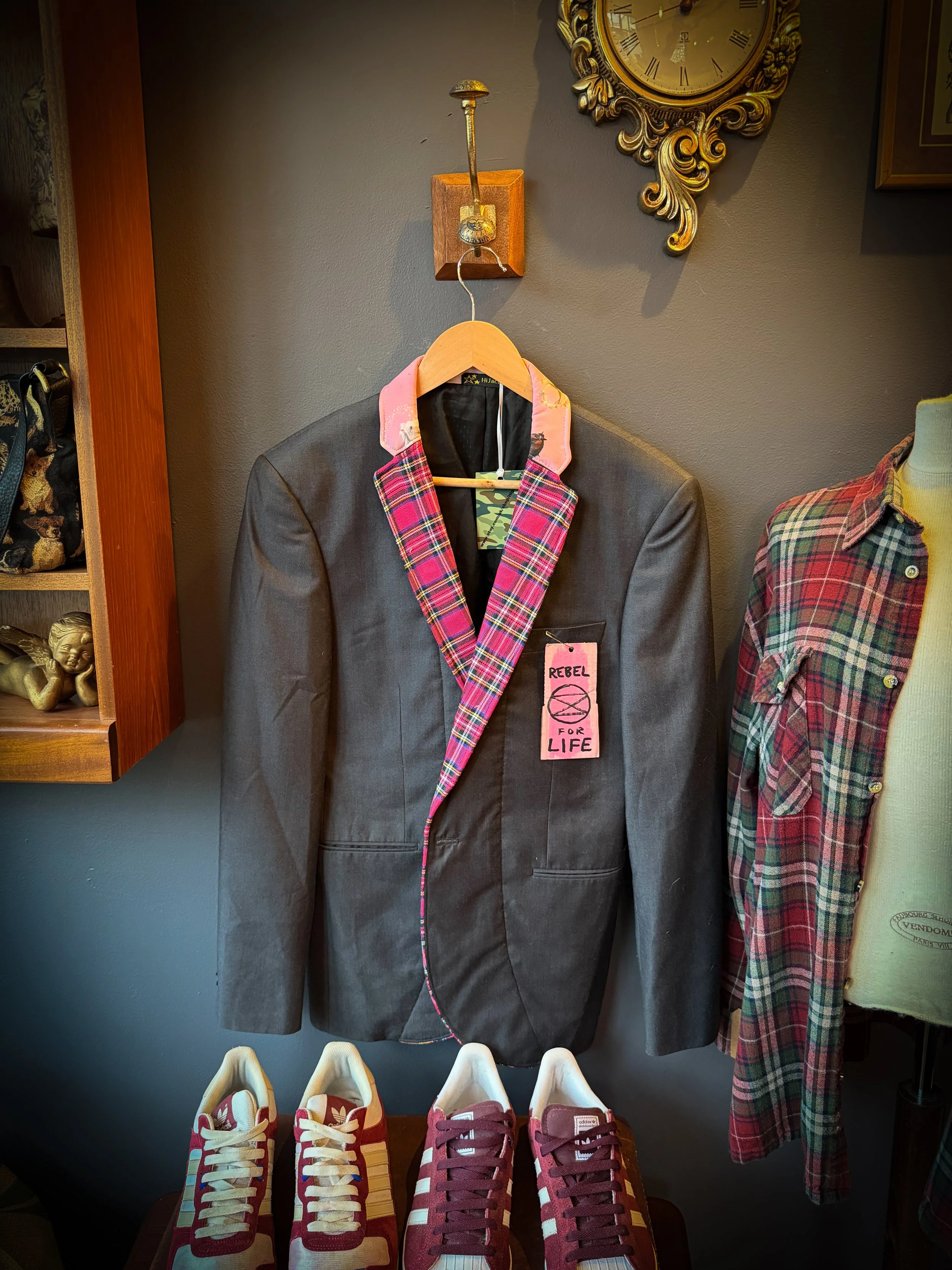 A clothing display featuring a black blazer with pink plaid lapels hanging on a hook, with a pink tag reading 'Rebel for Life'; four red-and-white sneakers with a pink Adidas logo are placed below.