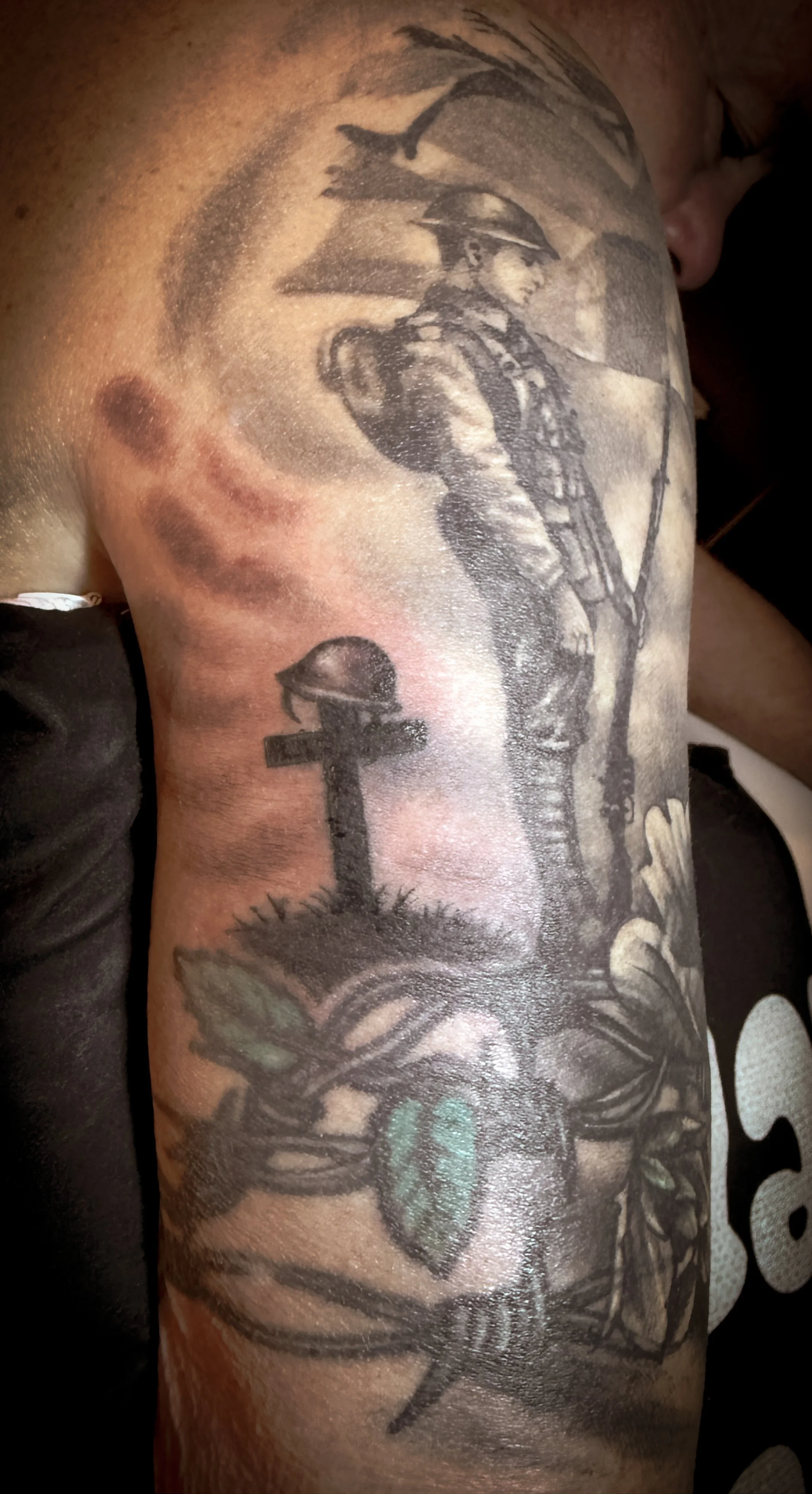 Black and gray tattoo of a soldier with a rifle and helmet, a cross with a helmet on top, and a heart with a banner and leaves.