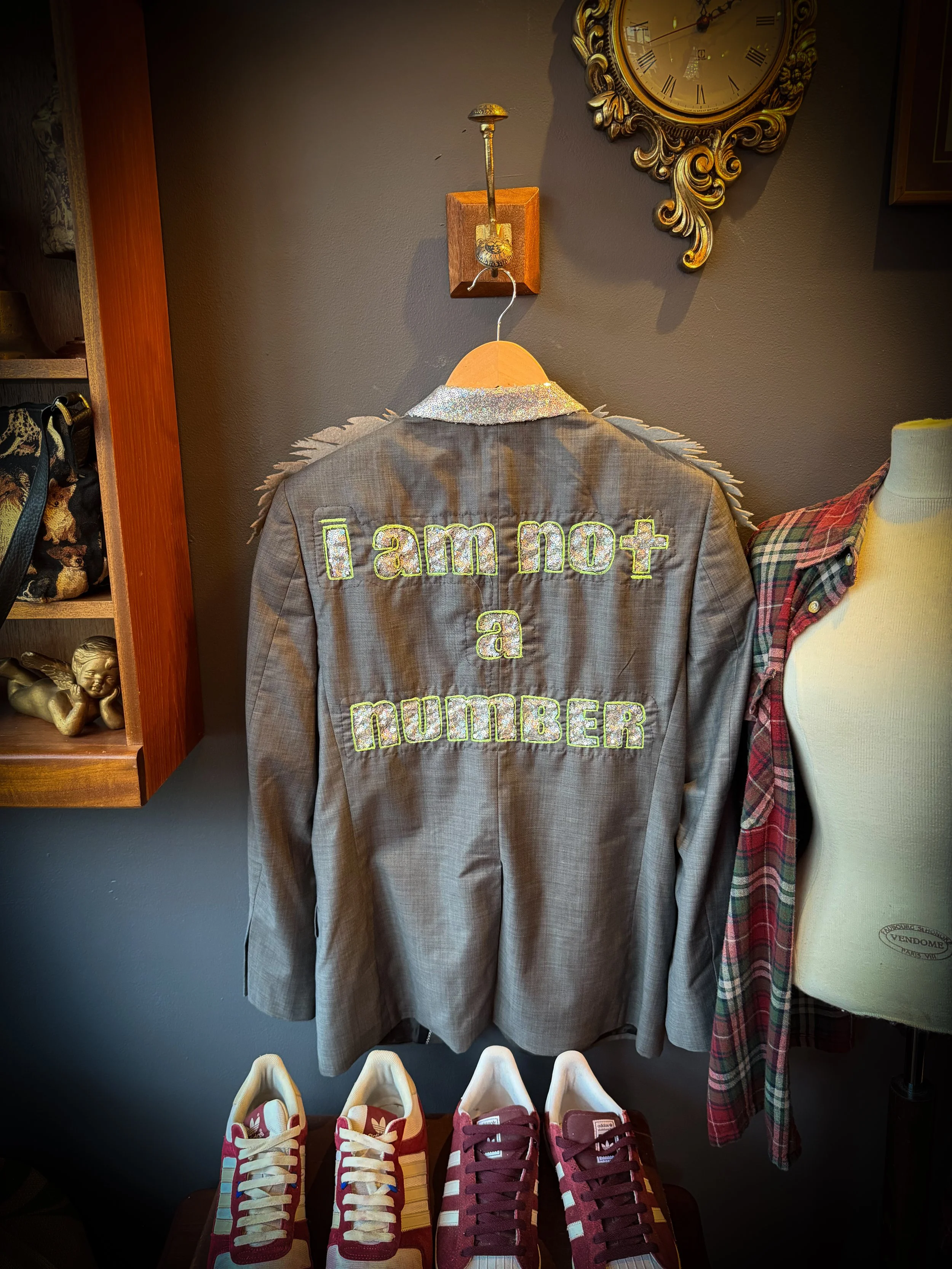Gray blazer with the phrase "I am not a number" embroidered on the back, hanging on a wooden hanger, with a clock and other objects on the wall behind, and a pair of red and beige sneakers below.