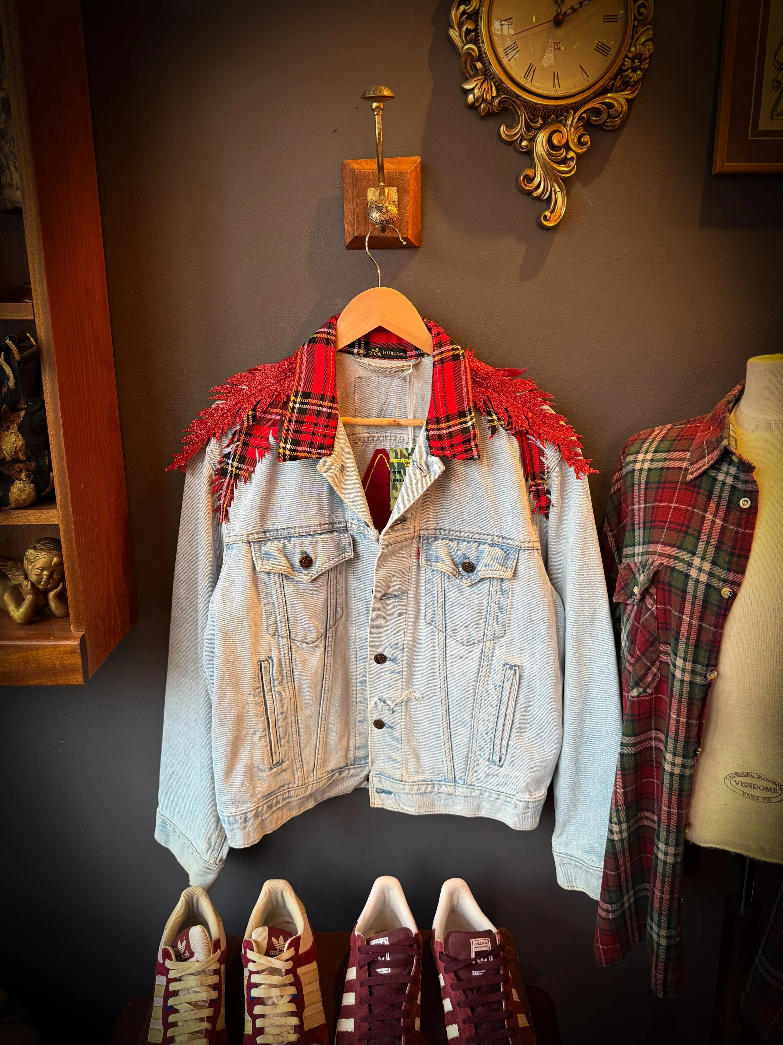 A denim jacket with red plaid and fringed shoulder accents hanging on a wooden hanger. There are three pairs of sneakers on a table below the jacket, and a vintage clock on the wall behind with a decorative frame.