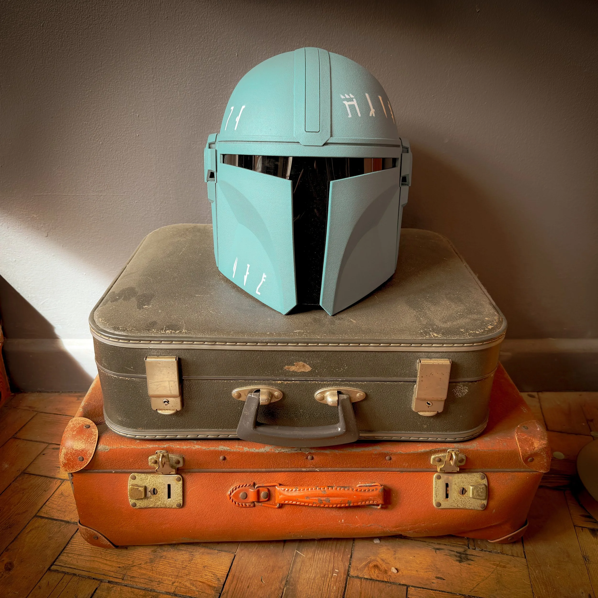 A Star Wars-themed Mandalorian helmet placed on two stacked vintage suitcases on a wooden floor near a wall.