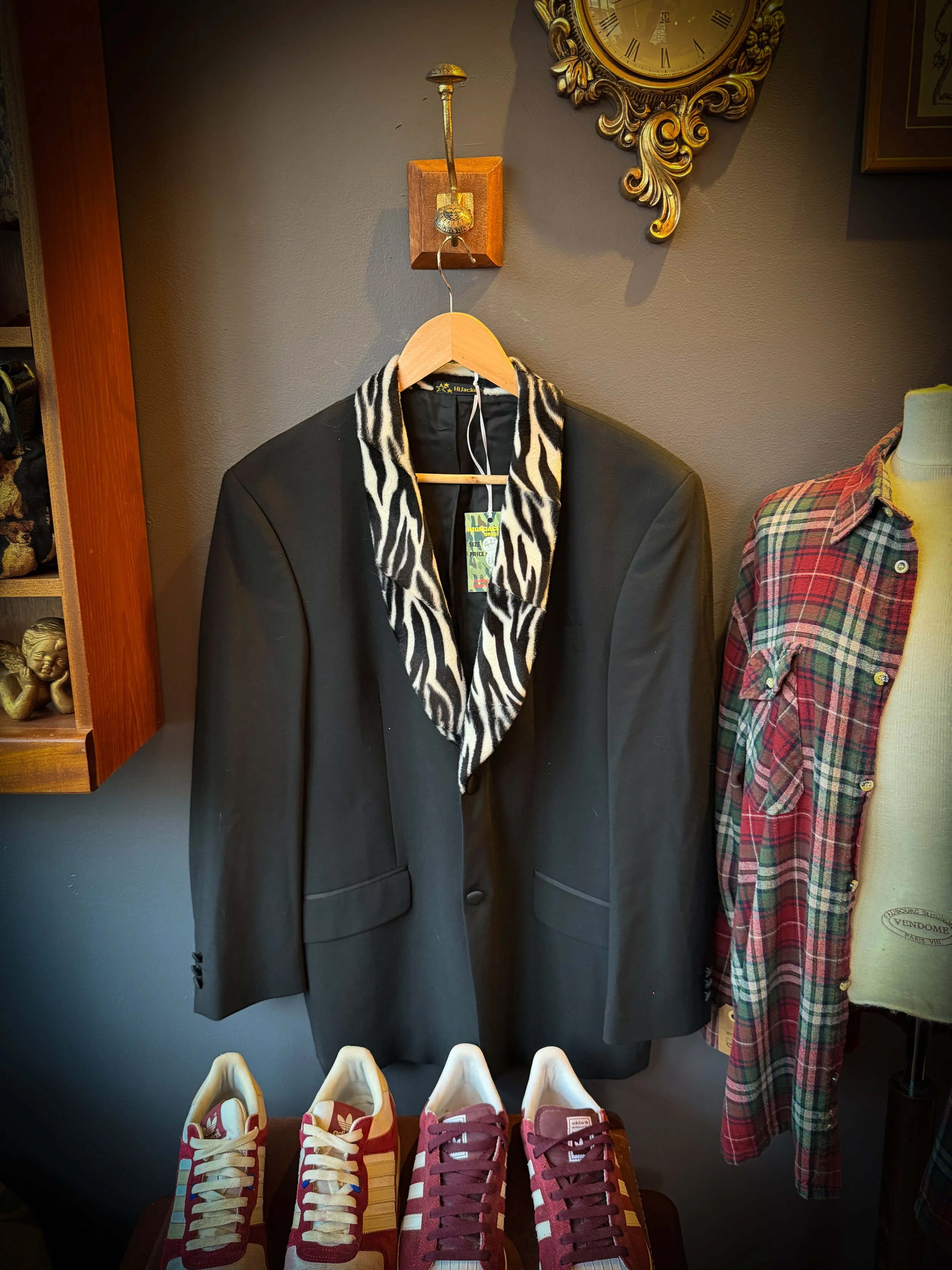 Black blazer with a zebra print collar hanging on a wooden hanger, displayed on a wall next to a mannequin dressed in a plaid shirt, and a row of red and white sneakers on a table below.