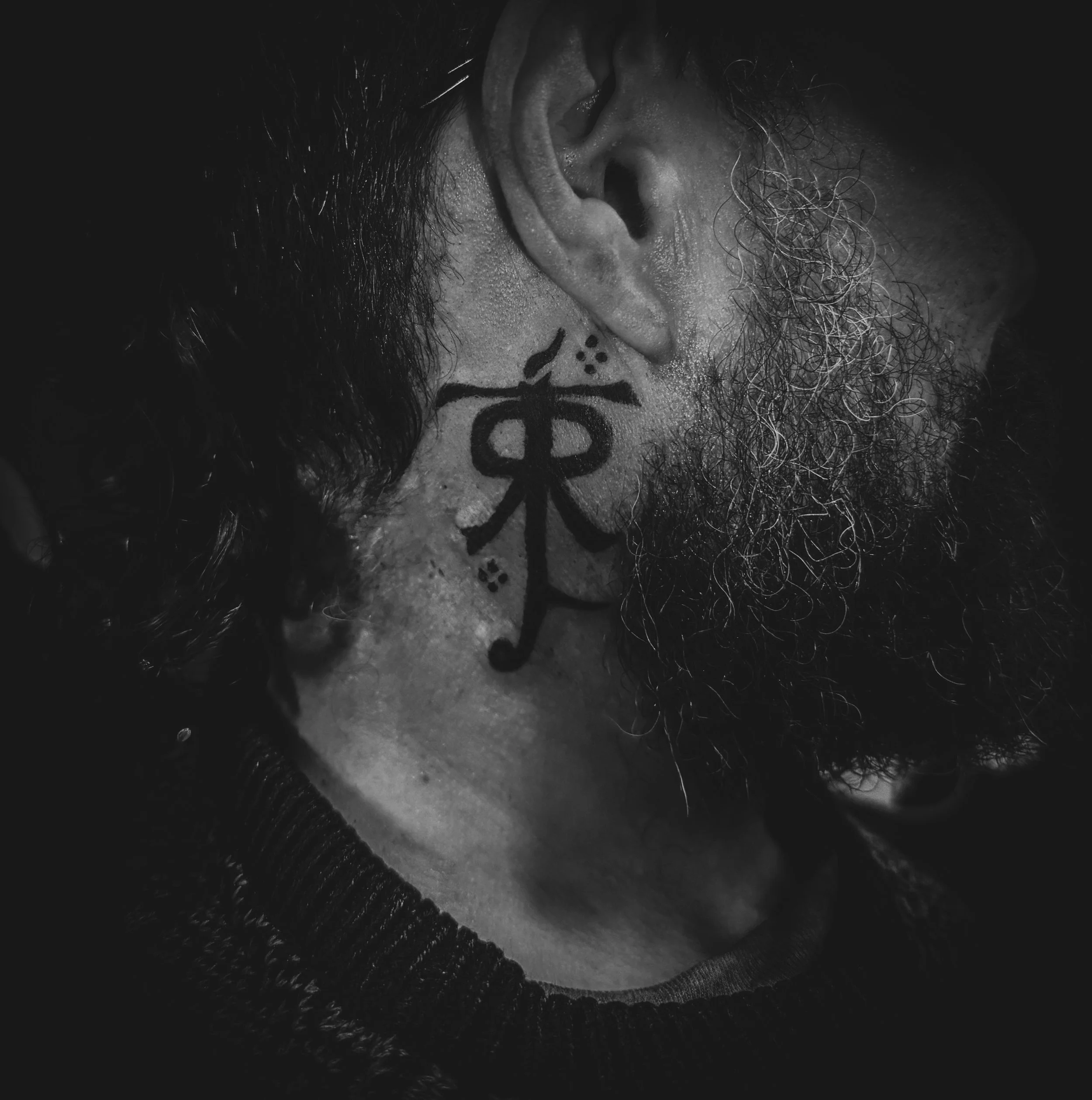 Close-up of a man's face with a beard, showing a tattoo on his neck, with his ear partially visible, in black and white.