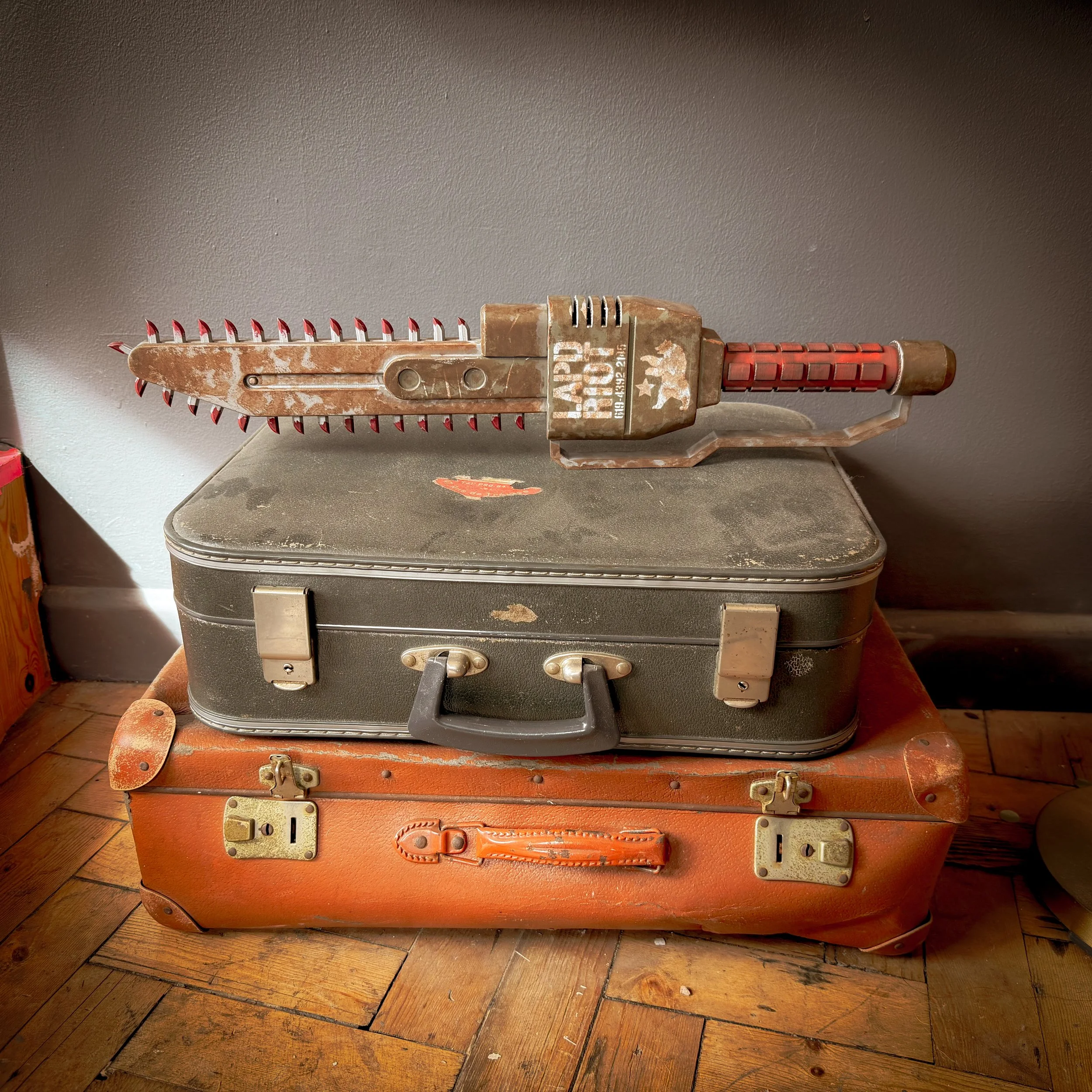 A vintage chain saw placed on top of two stacked suitcases, one black and one orange, on a wooden floor against a plain wall.