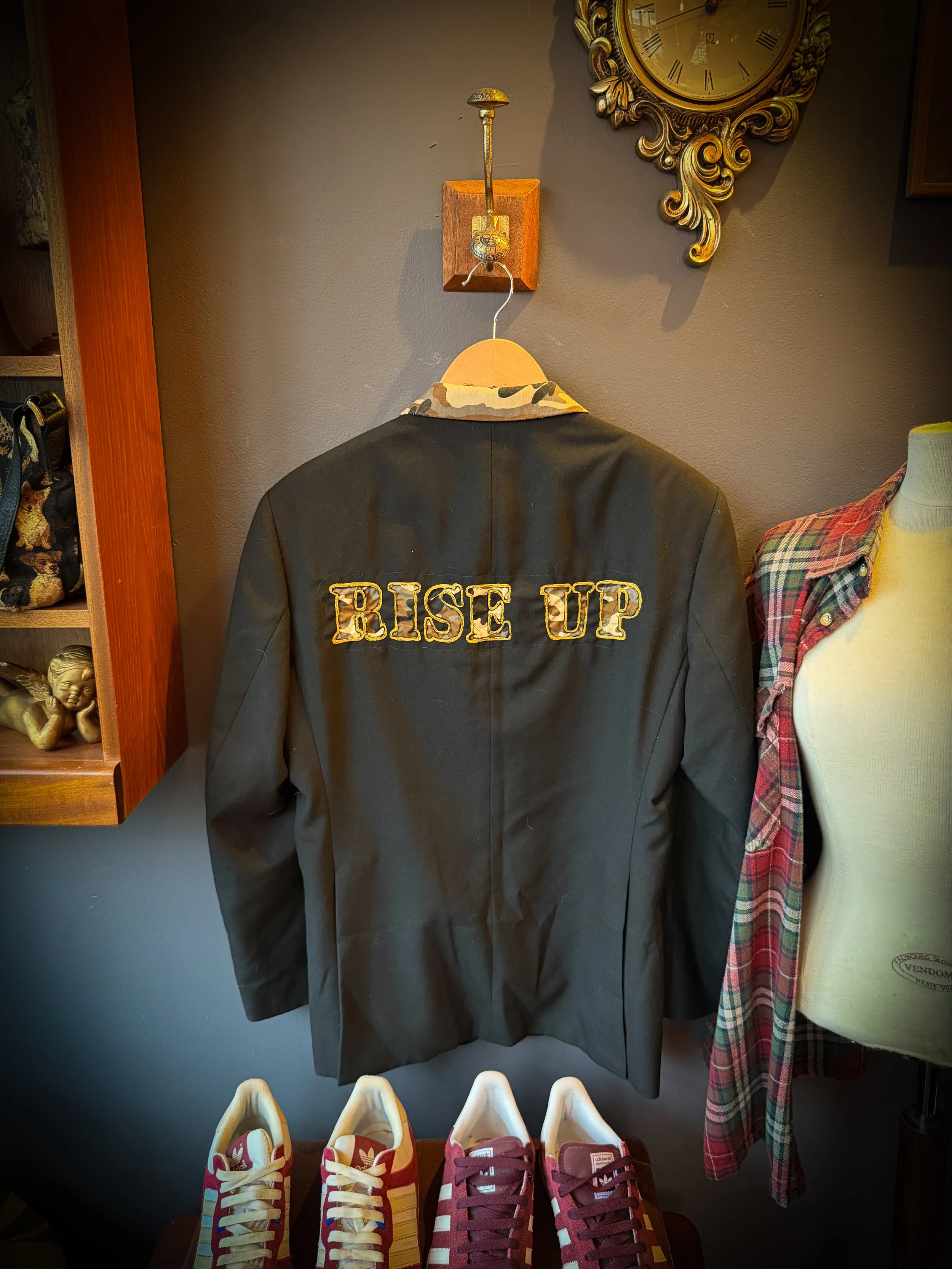 A black jacket with 'RISE UP' embroidered on the back, hanging on a wooden hanger, with a plaid shirt partially visible to the right, and a pair of red and white sneakers in the foreground, on a table.