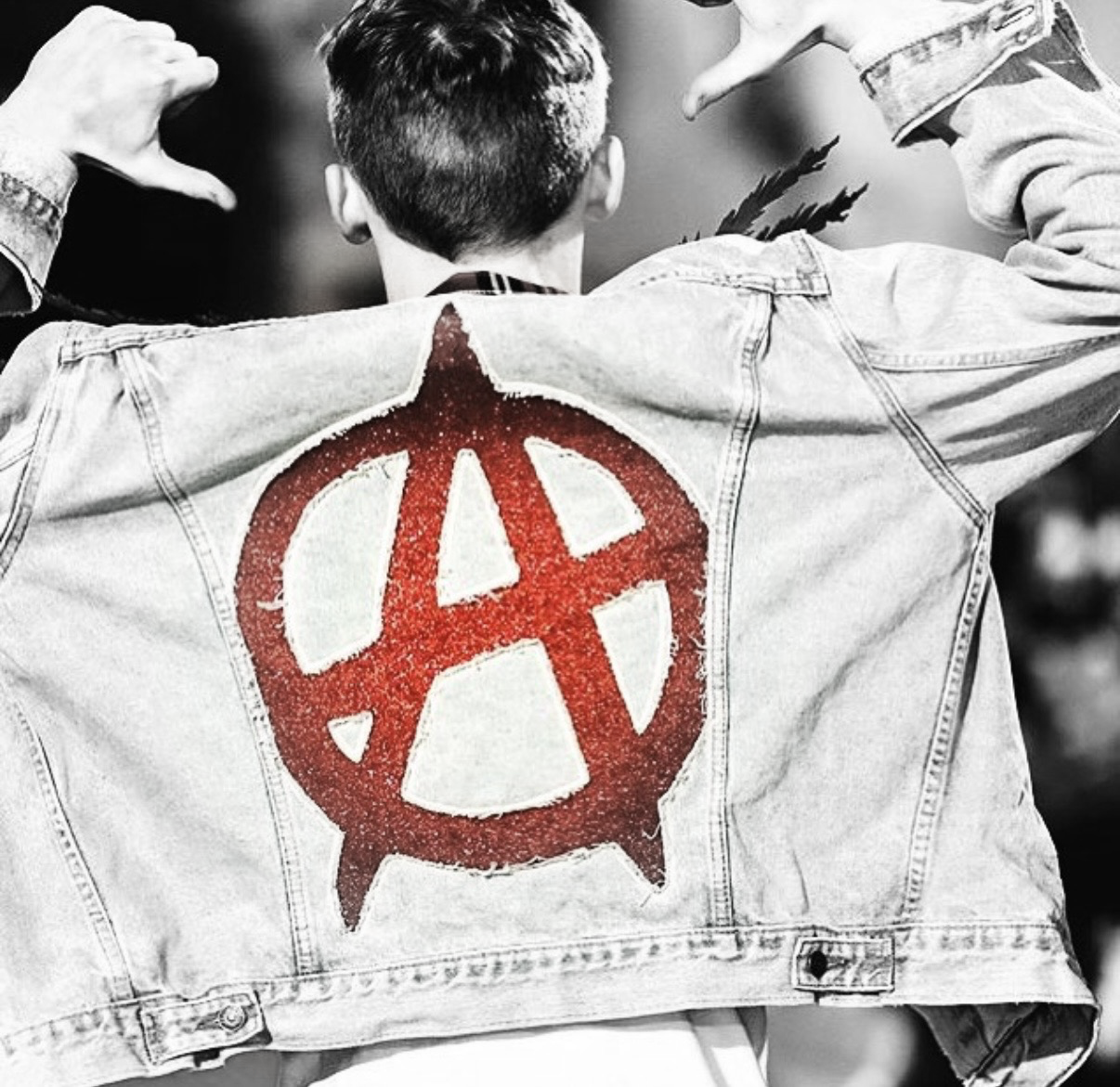 Back of a person wearing a denim jacket with a large red anarchist symbol painted on it, facing away and making a hand gesture with both hands.