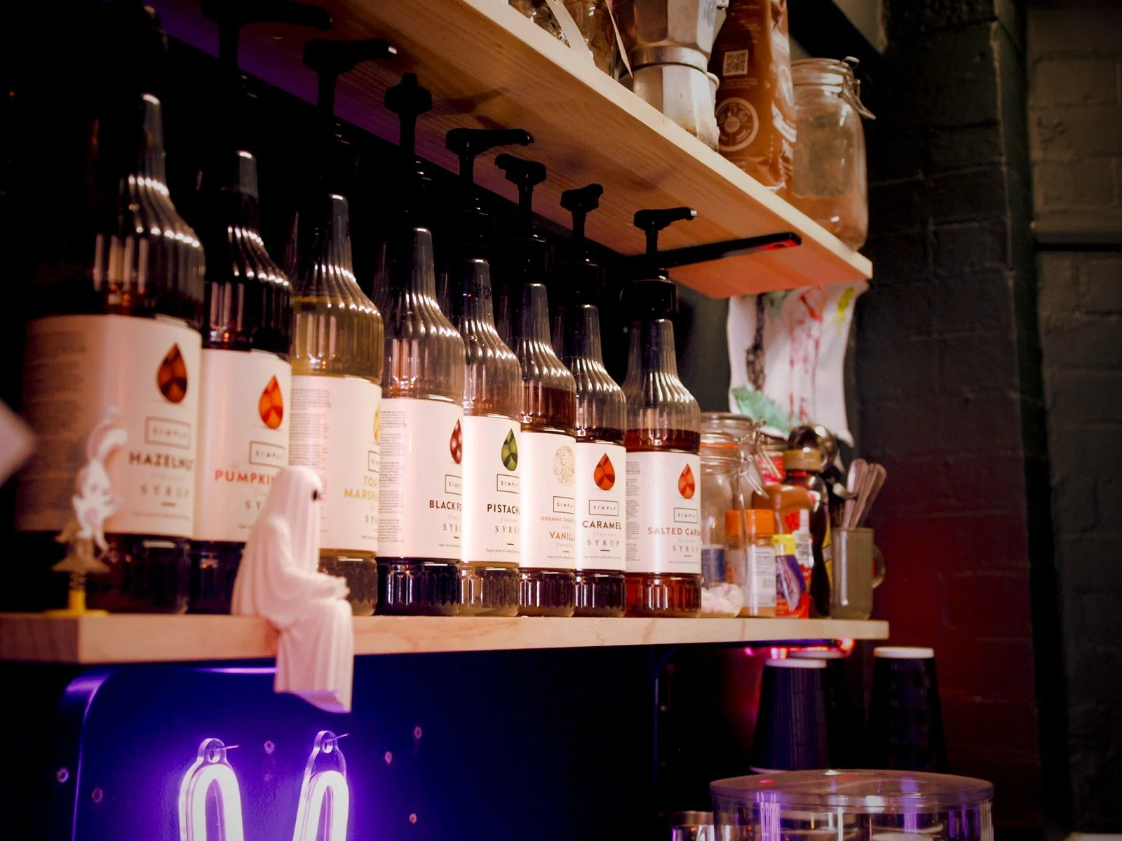 A shelf holding various bottles of flavored syrups in a cafe or coffee shop, with labels indicating flavors like hazelnut, pumpkin, and salted caramel, and other jars and supplies in the background.