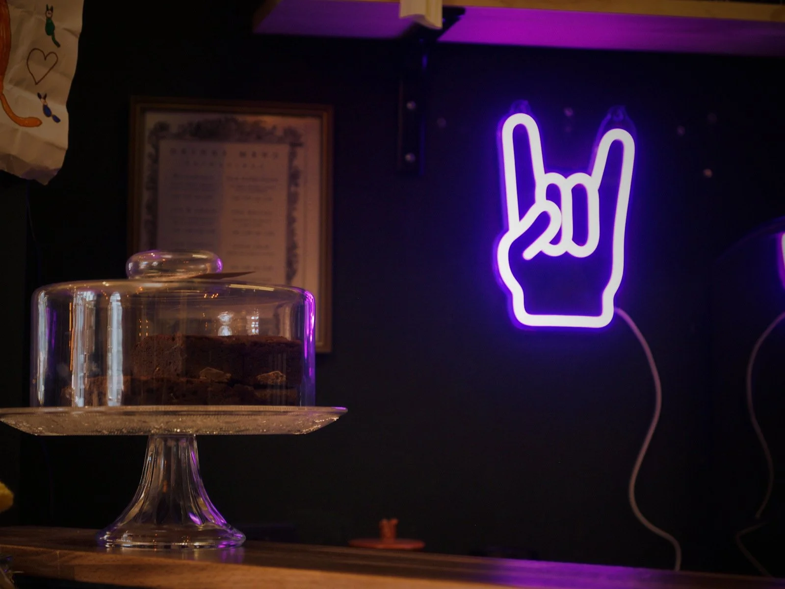 A glass cake stand with a dome lid containing a chocolate cake, set on a wooden surface. In the background, a purple neon sign shaped like a hand making the 'rock on' gesture with the number 20 inside the fingers, and a framed document on a dark wall.