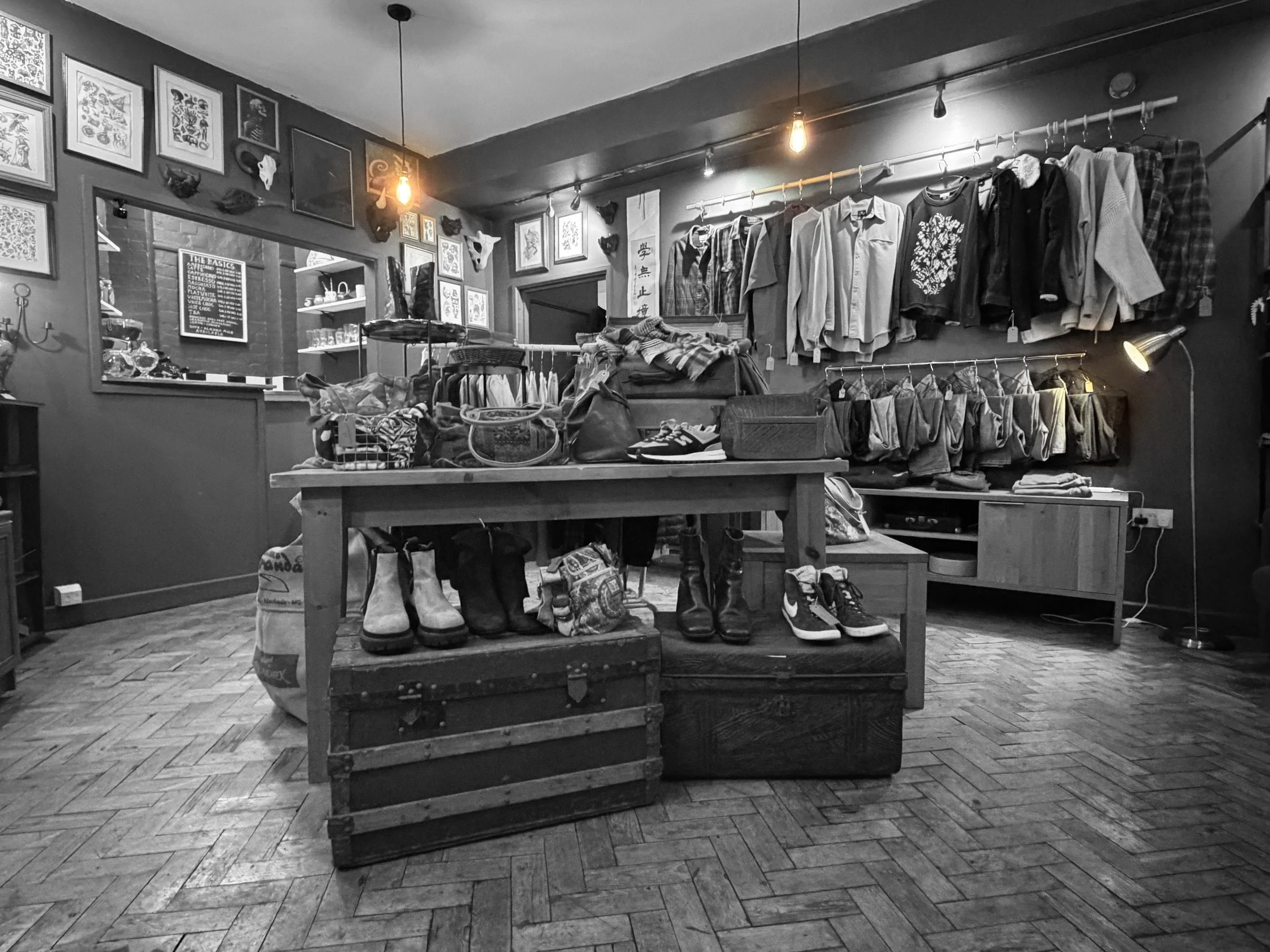 A black and white photo of a clothing store with various shirts, shoes, and accessories displayed on tables and shelves, and framed artwork on the walls.