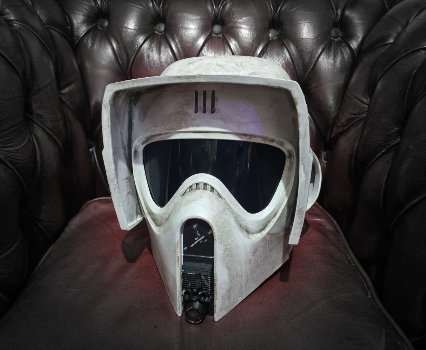 A vintage, weathered white helmet with a visor and breathing apparatus, resting on a dark leather couch.