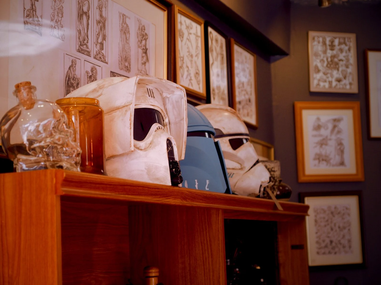 A wooden shelf displays three Stormtrooper helmets, two white and one blue, along with two glass bottles and framed artwork on the wall behind.
