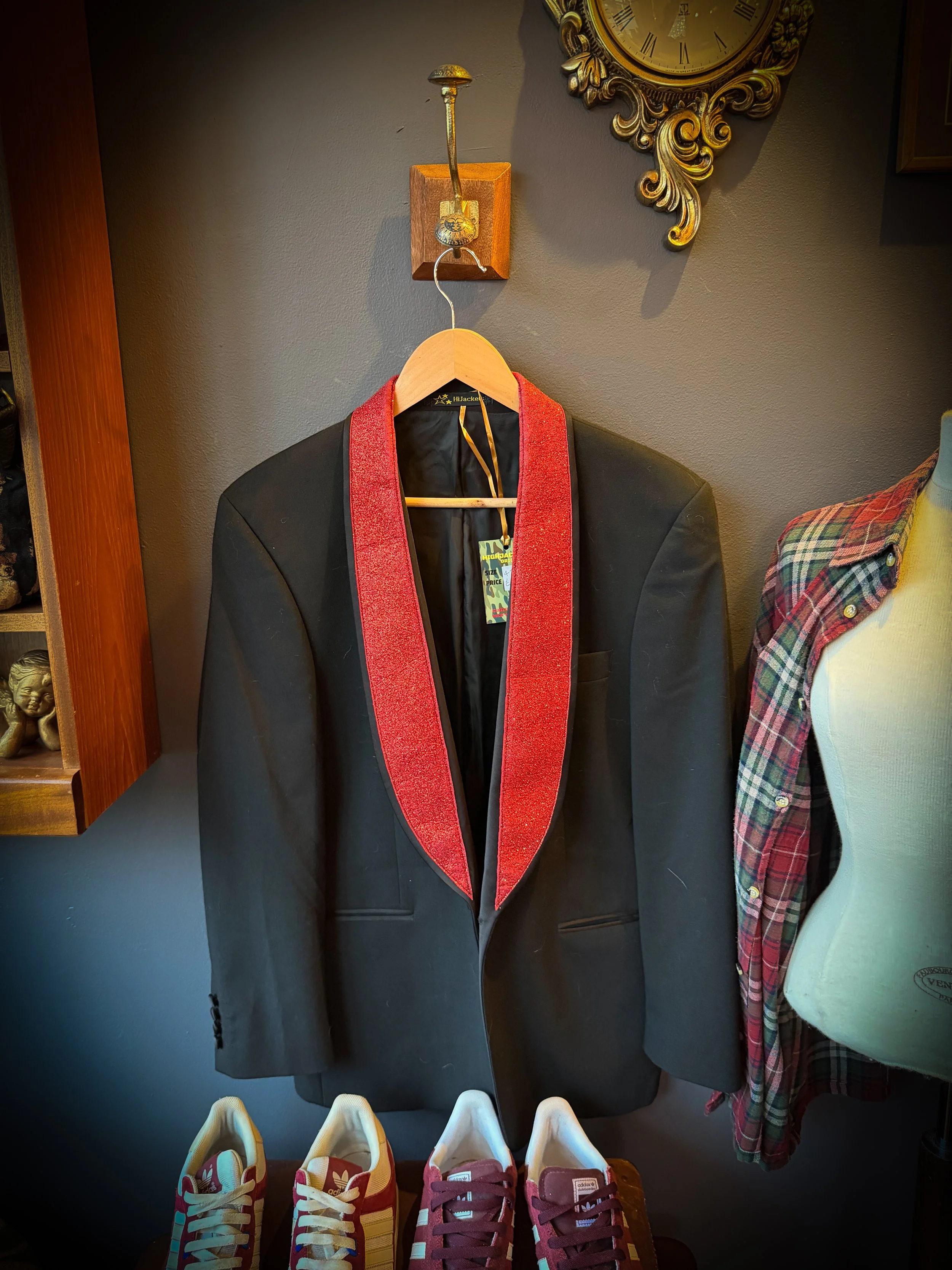 A black blazer with a red sparkly lapel hanging on a wooden hanger on a wall. Part of a mannequin wearing a plaid shirt is visible on the right. Three pairs of colorful sneakers are in front of the blazer.