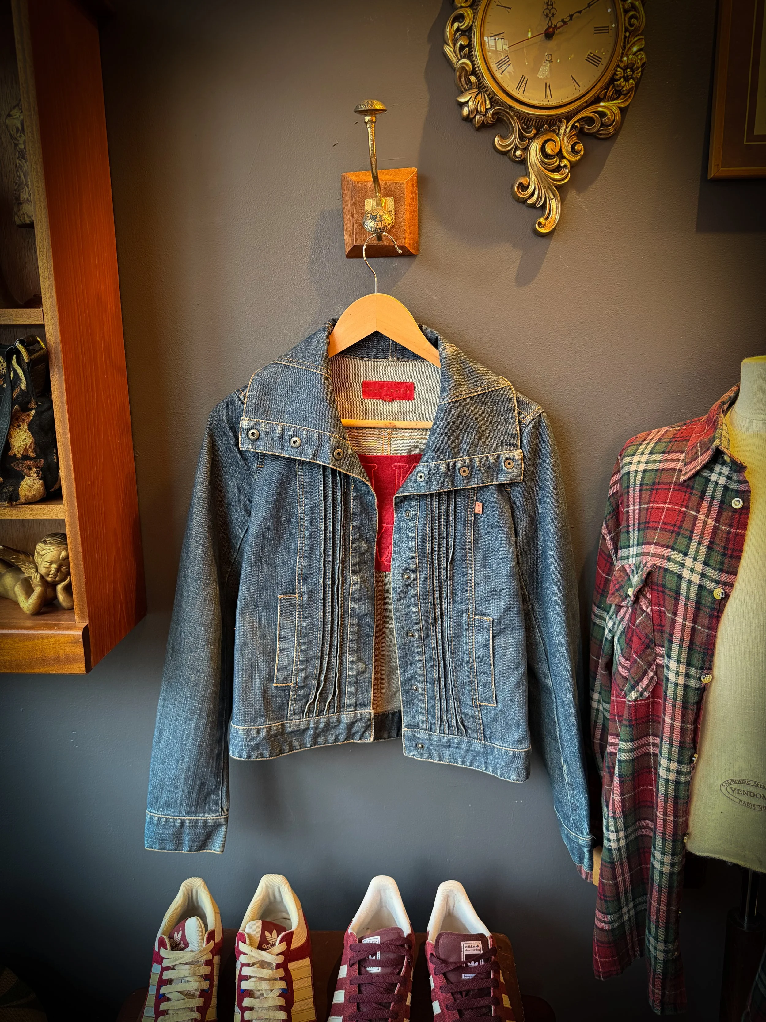 A denim jacket hanging on a hanger on a gray wall, placed below an ornate gold clock. To the right, part of a plaid shirt on a mannequin. Shoes are displayed on a table below the jacket.