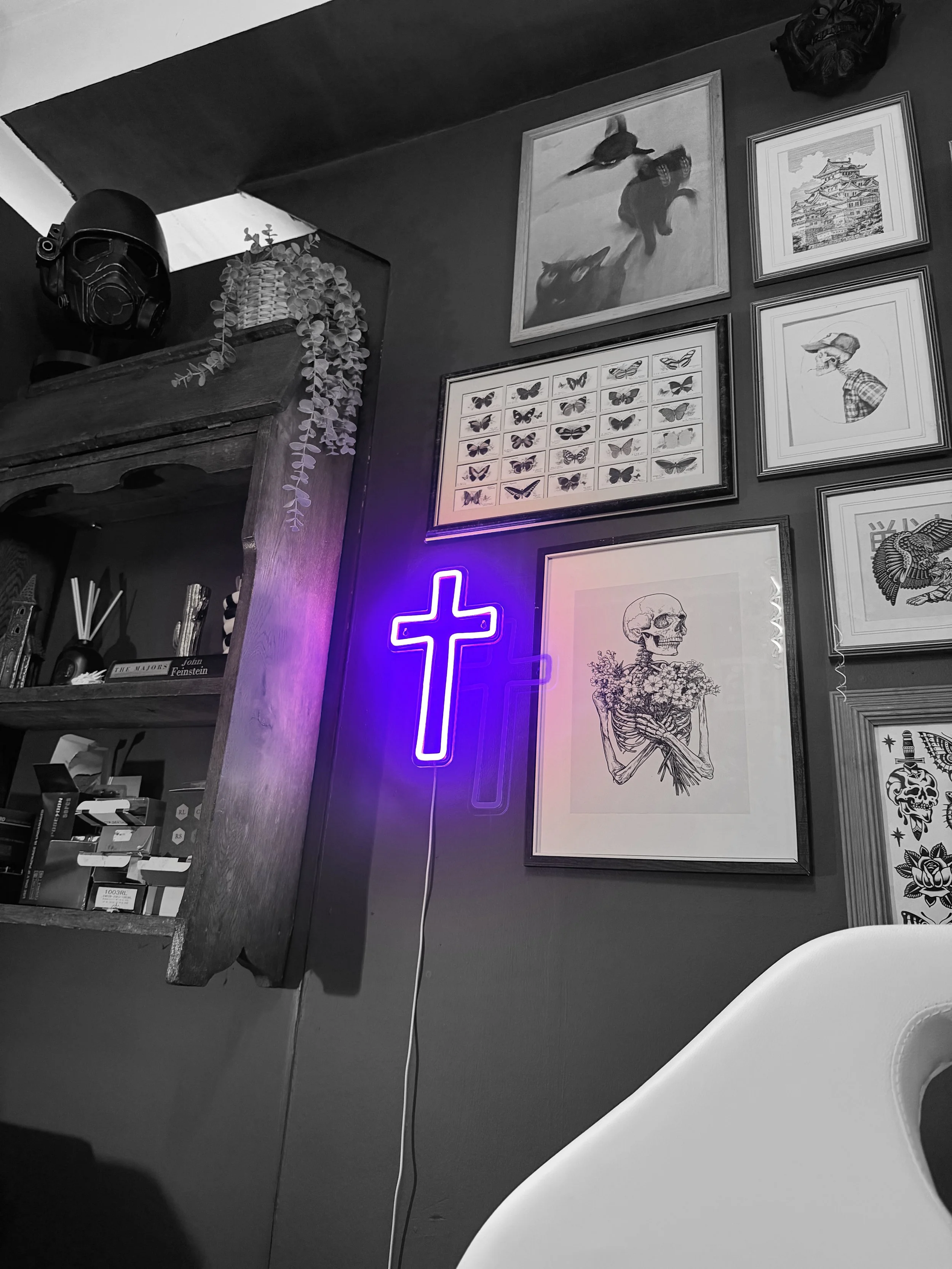 Black-and-white photo of a wall decorated with framed pictures, including a purple neon cross sign, a picture of a skeleton with flowers, and other artwork such as animals, architecture, and portraits, with a wooden shelf and a black helmet on top.