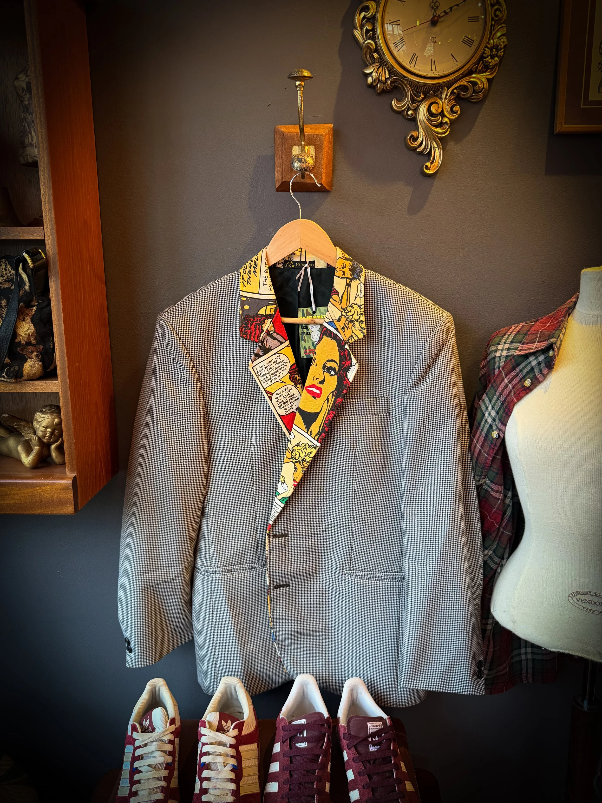 A gray checkered blazer with a colorful comic book style lining on a wooden hanger, positioned before a wall clock and a mannequin torso with a plaid shirt.