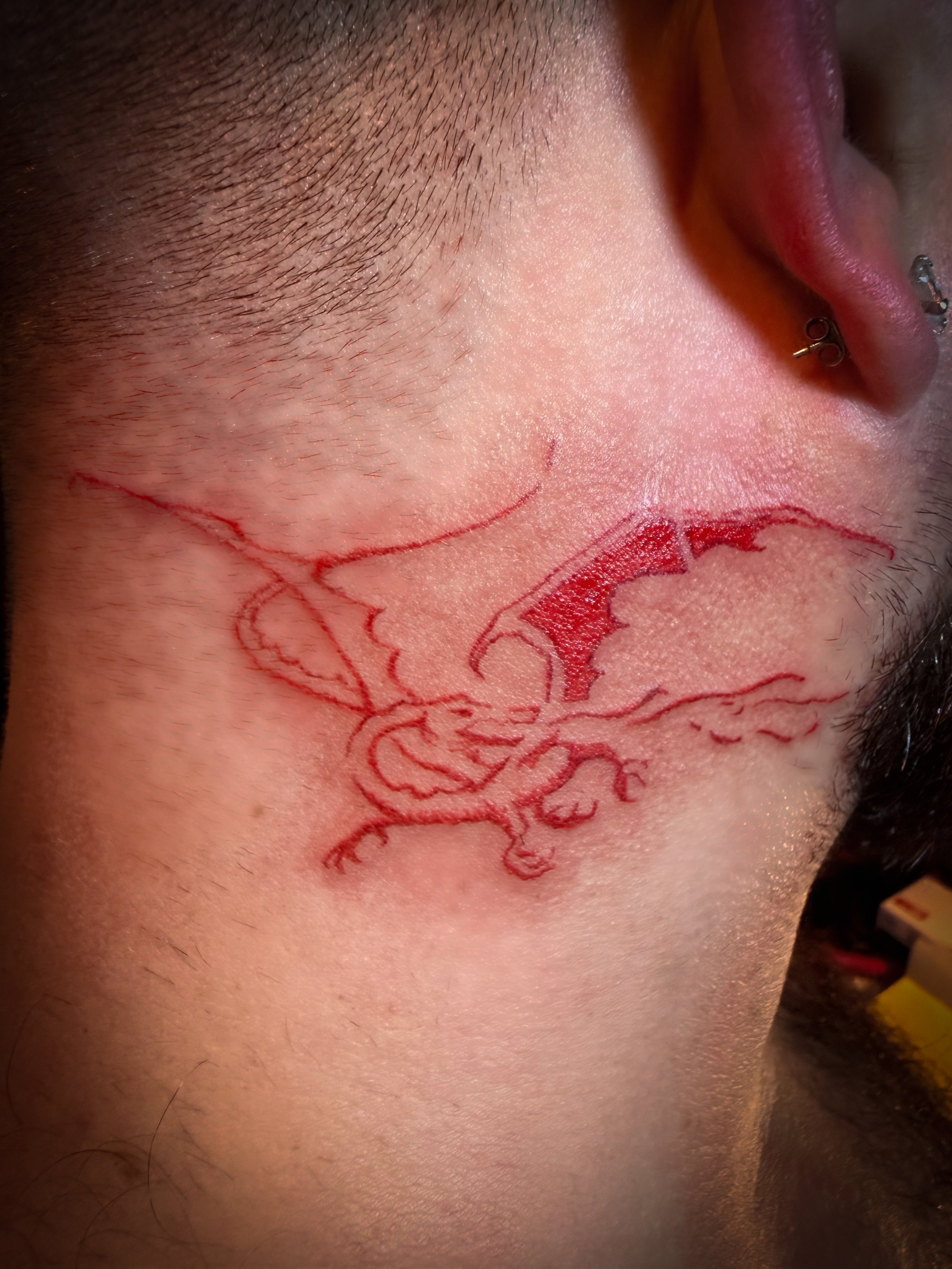 Close-up of a person's face showing freshly tattooed red outline of a dragon on their cheek, with some redness and swelling.