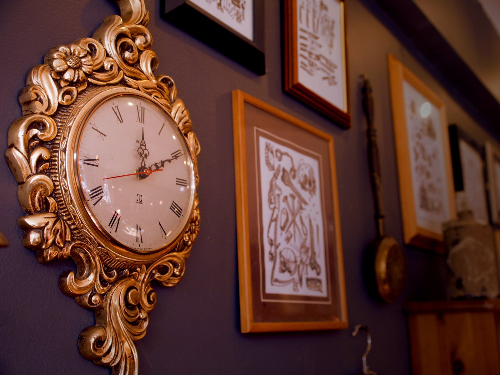 Decorative clock with Roman numerals on a dark wall surrounded by framed artwork and antique items.