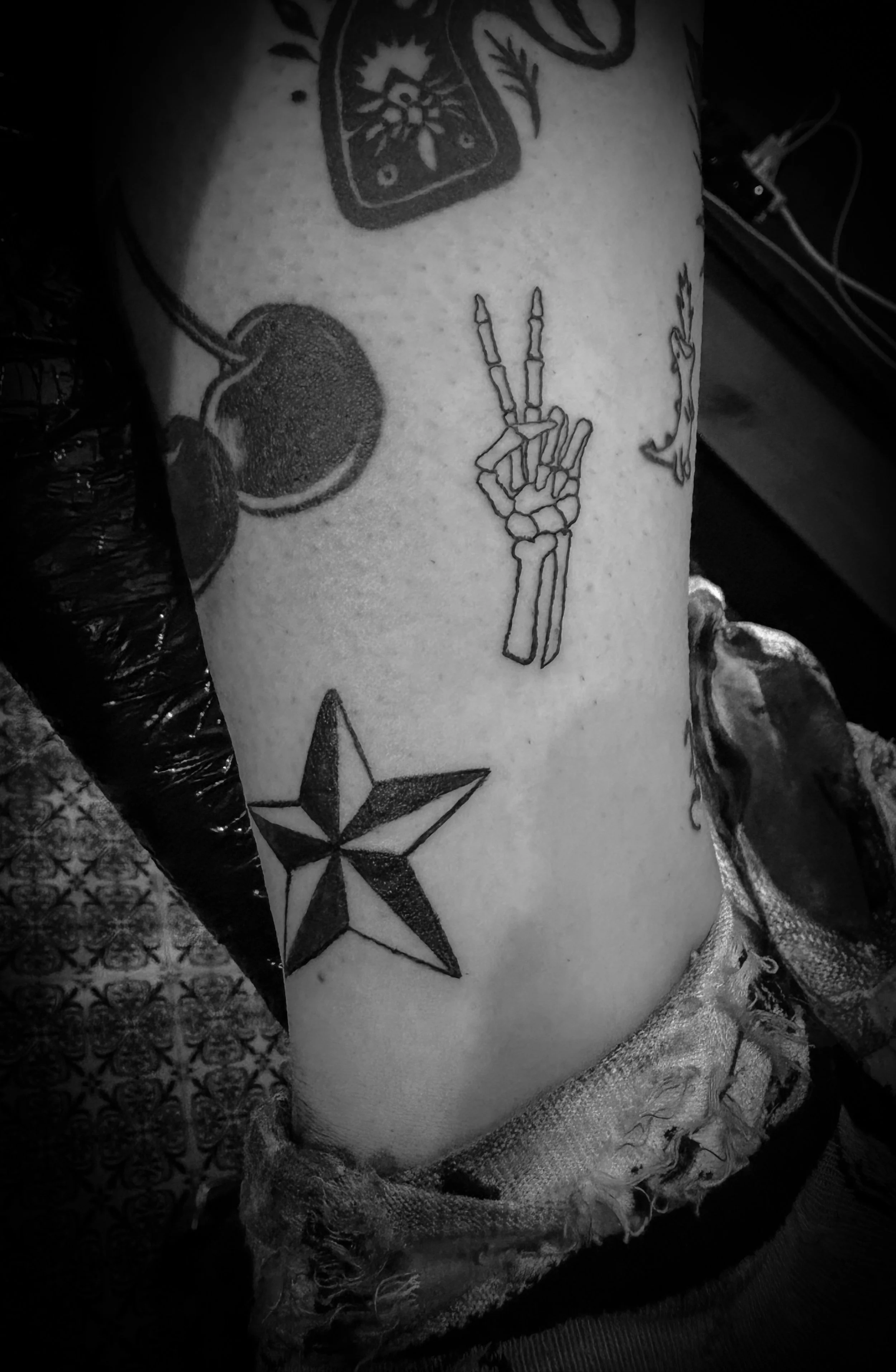 Black and white photo of a person's arm with several tattoos, including cherries, a star, a skeleton hand making a peace sign, and other designs.